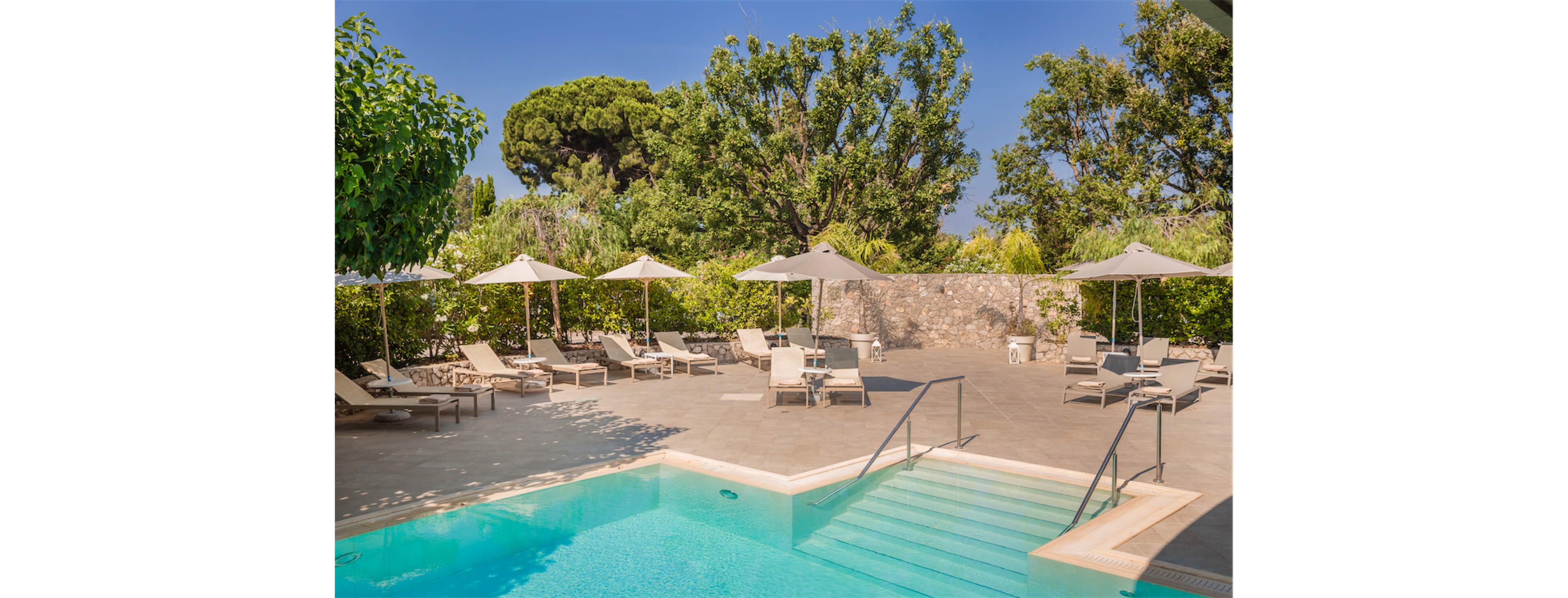 An inviting outdoor swimming pool sits in the foreground with clear turquoise water. Surrounding the pool is a spacious tiled deck lined with sun loungers and beige umbrellas. Lush green trees and shrubs create a private, tranquil atmosphere around the area. A stone wall in the background adds rustic charm under bright, sunny skies.