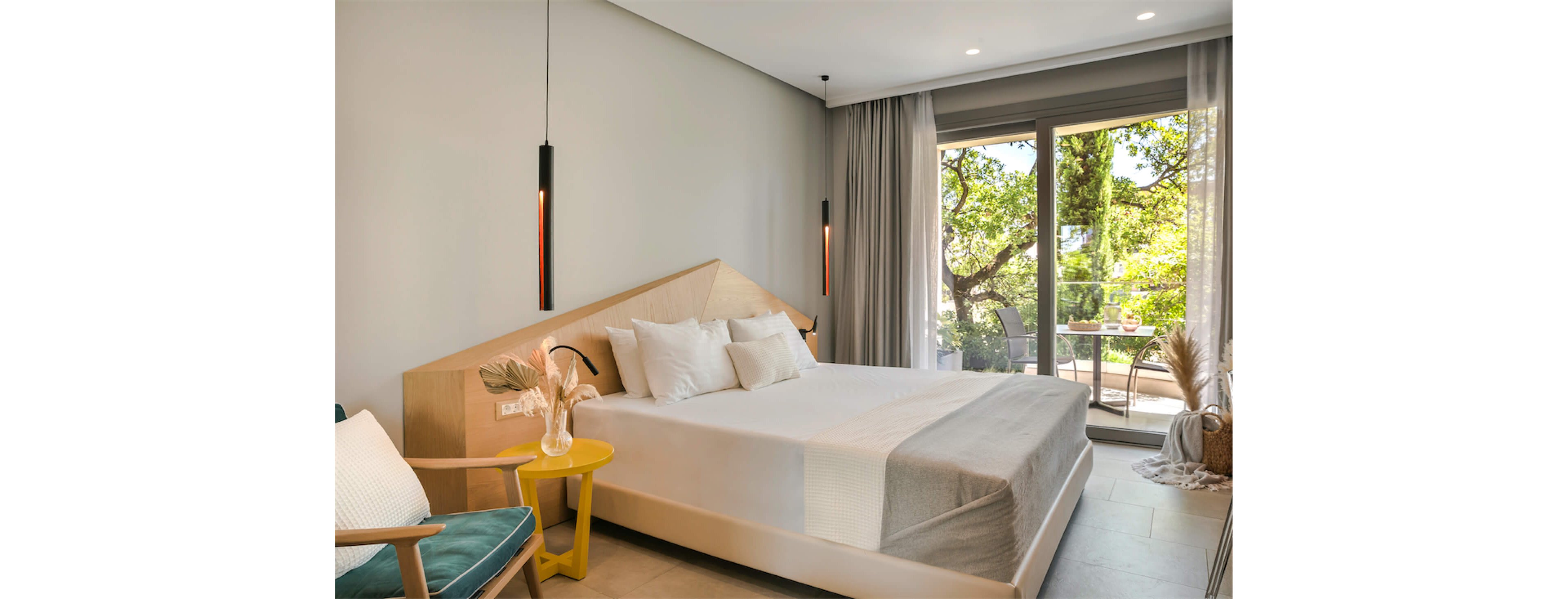 A contemporary bedroom features a large neatly made bed with white linens and neutral blankets. Minimalist pendant lights hang on either side of the wooden headboard, illuminating the space softly. A yellow side table, accent chair, and decorative dried plants add warmth and texture to the room. Floor-to-ceiling glass doors open to a terrace with outdoor seating and lush green trees visible outside.
