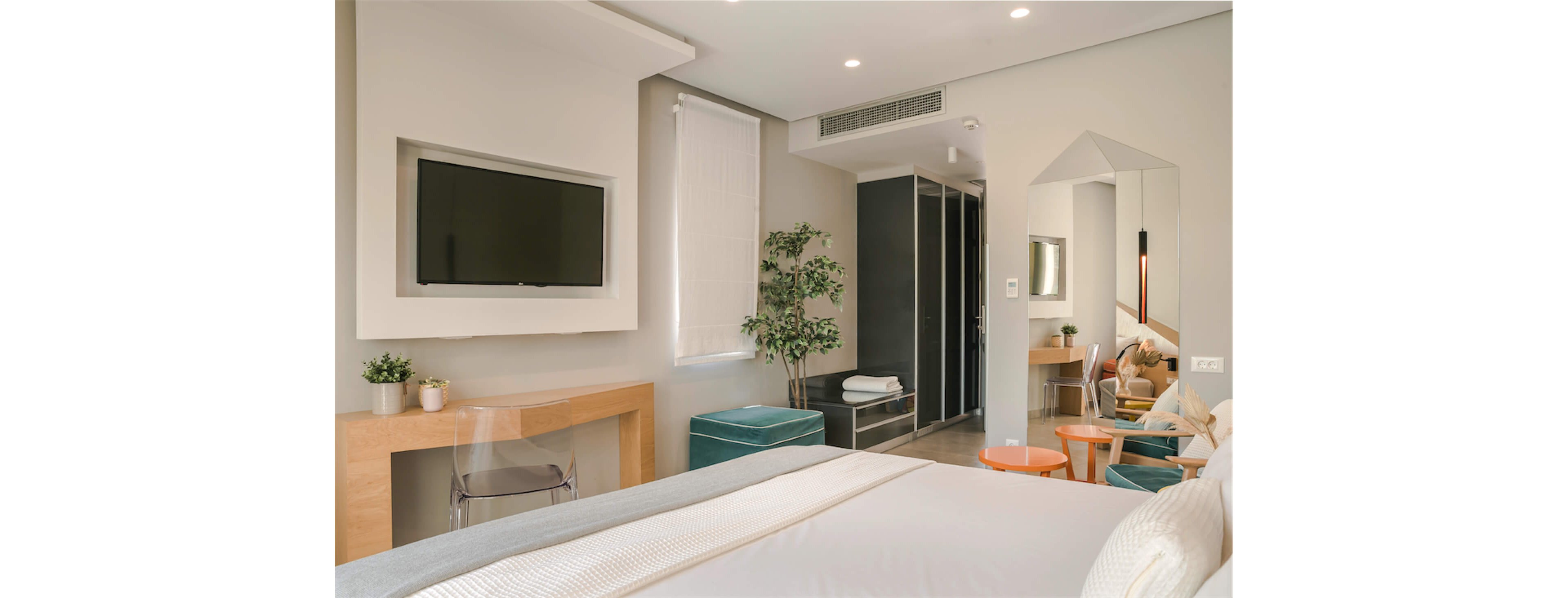 A contemporary hotel bedroom features a neatly made bed in the foreground. A wall-mounted flat-screen television and light wood desk sit opposite the bed, with a transparent chair tucked underneath. Soft neutral tones, recessed ceiling lights, and a potted plant create a calm, minimalist atmosphere. In the background, a dressing area with mirrors, stools, and a wardrobe adds functionality to the stylish space.