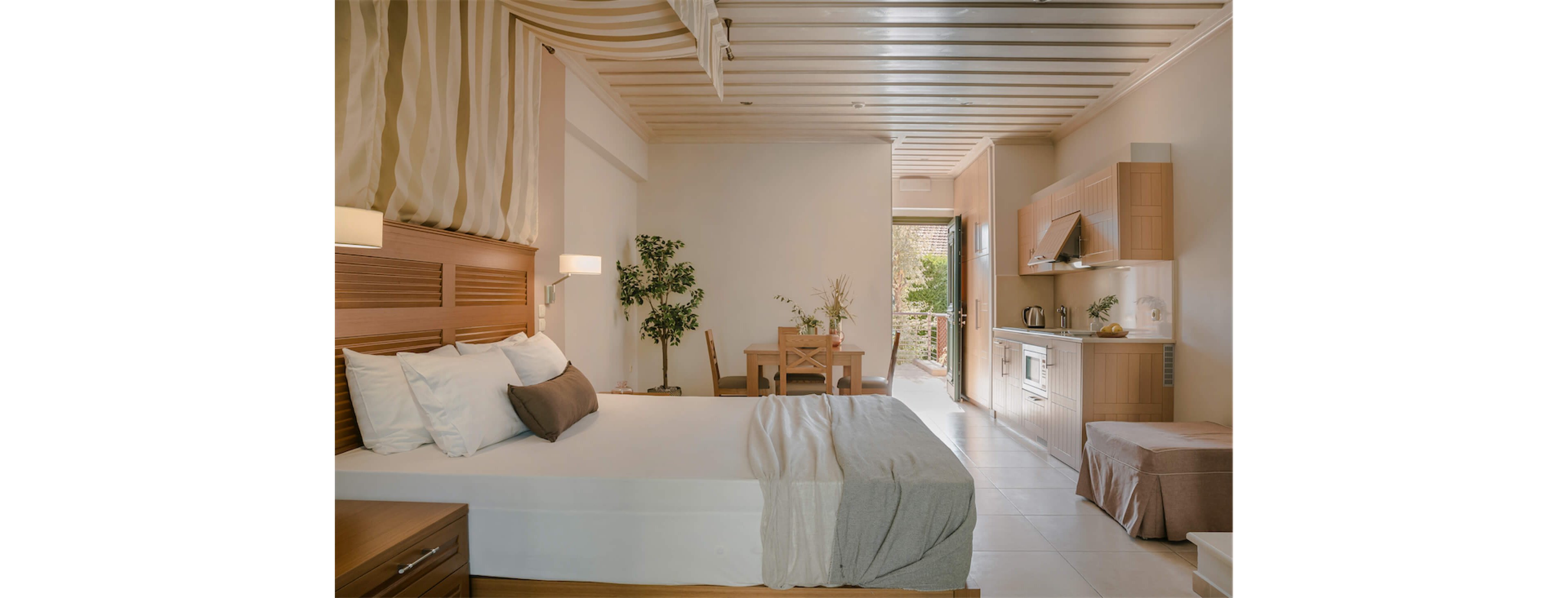A neatly made double bed with neutral linens sits in a bright studio room. The space extends to a small dining table and chairs, with potted plants adding greenery. A compact kitchenette with cabinets, stove, and appliances lines the right wall. Soft, warm lighting and beige tones create a calm, inviting atmosphere, with daylight visible through the open doorway.