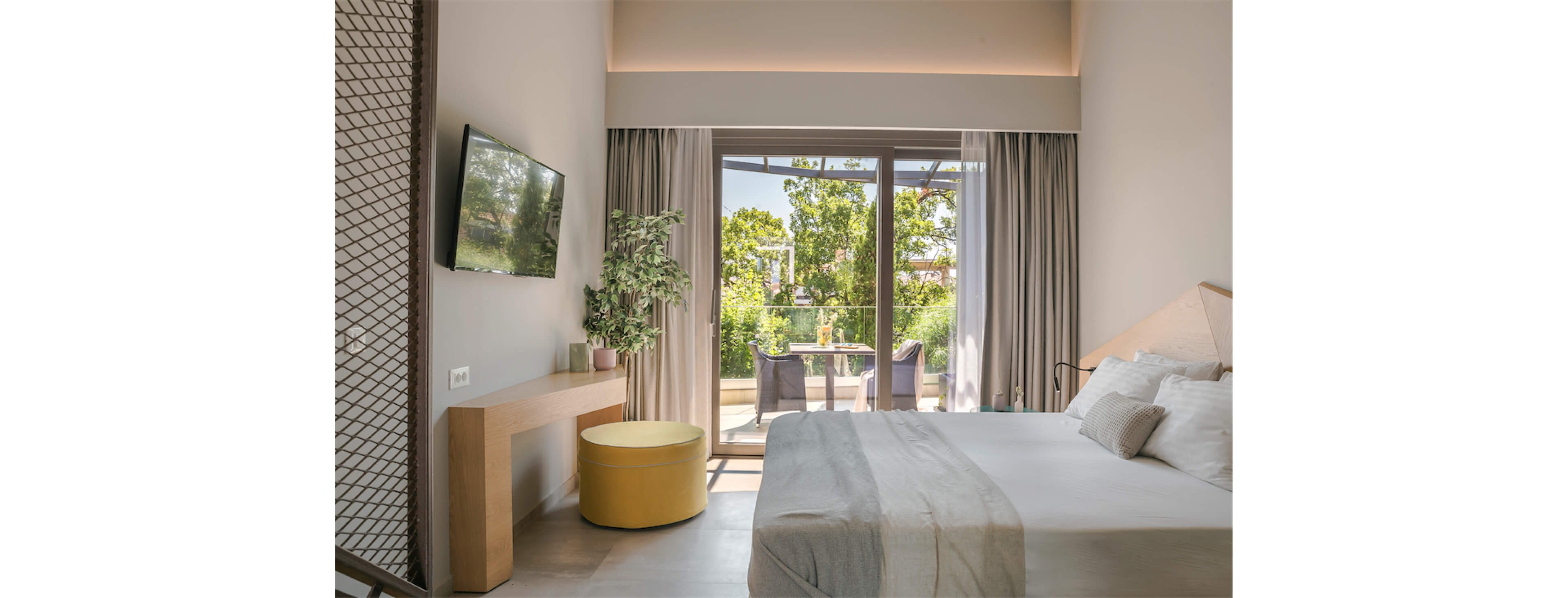 A contemporary hotel bedroom features a neatly made double bed with soft neutral linens and pillows. Floor-to-ceiling glass doors open onto a small balcony with chairs and a table overlooking lush green trees. A wall-mounted flat-screen TV, minimalist wooden console, and round yellow ottoman add functional style to the space. Soft natural light and muted colors create a calm, relaxing atmosphere ideal for rest or vacation stays.