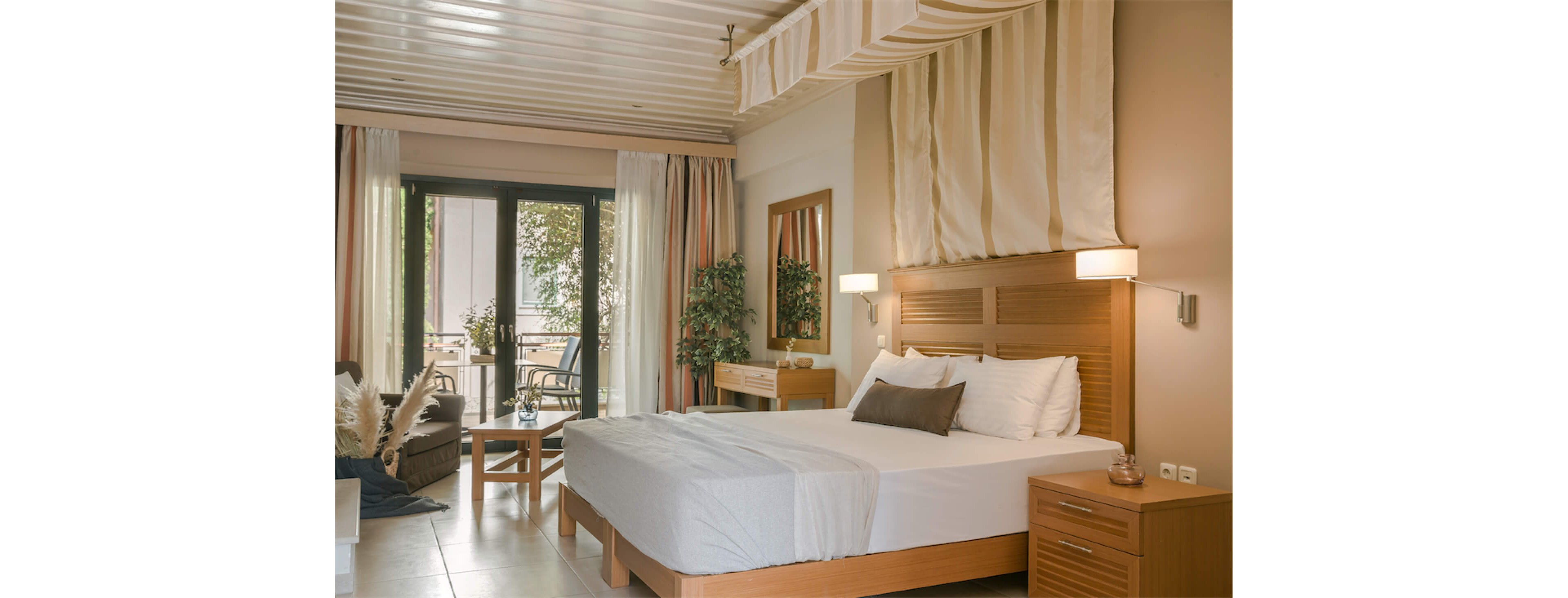 A spacious, modern bedroom features a large bed with crisp white linens and light wood furniture. Floor-to-ceiling curtains frame glass doors that open onto a balcony with outdoor seating. Soft neutral tones, indoor plants, and warm wall lights create a calm, inviting atmosphere. A small sitting area with an armchair and table adds comfort and functionality to the room.
