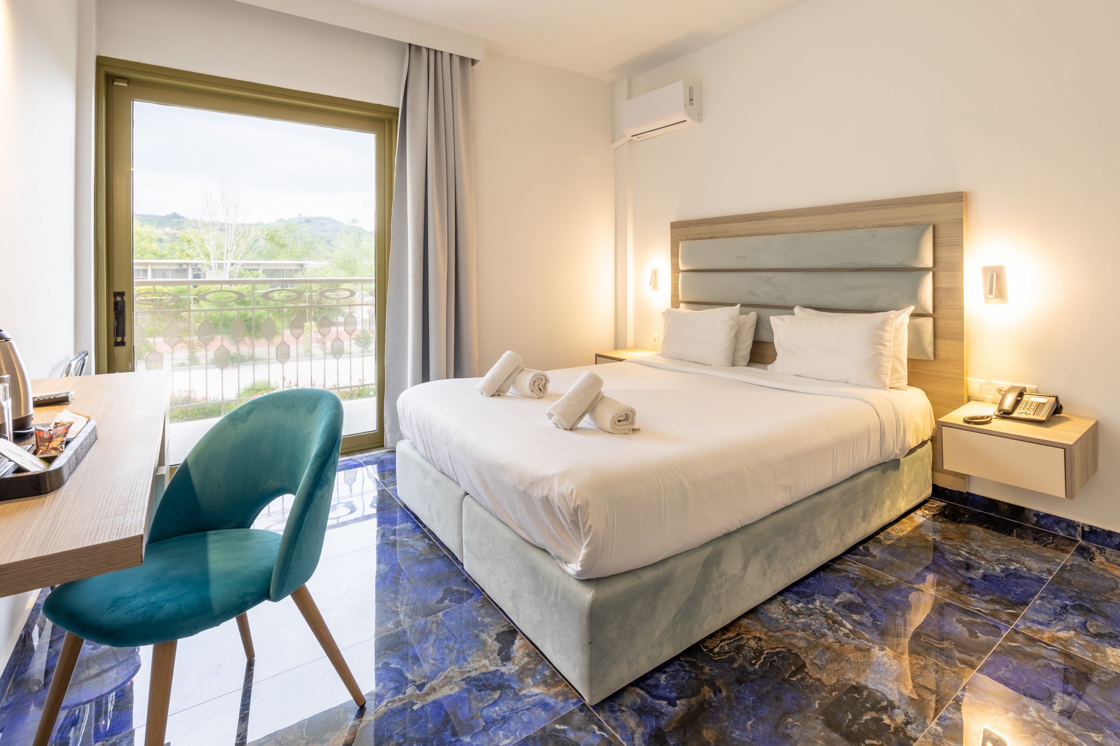 A contemporary hotel room features a large double bed with neatly folded towels placed on top. The space includes a small desk with a teal upholstered chair and a tray with a kettle and cups. Glossy blue marble-style flooring contrasts with the light walls and minimalist furniture. A sliding glass door opens to a balcony with a view of greenery and distant hills, creating an airy and inviting atmosphere.