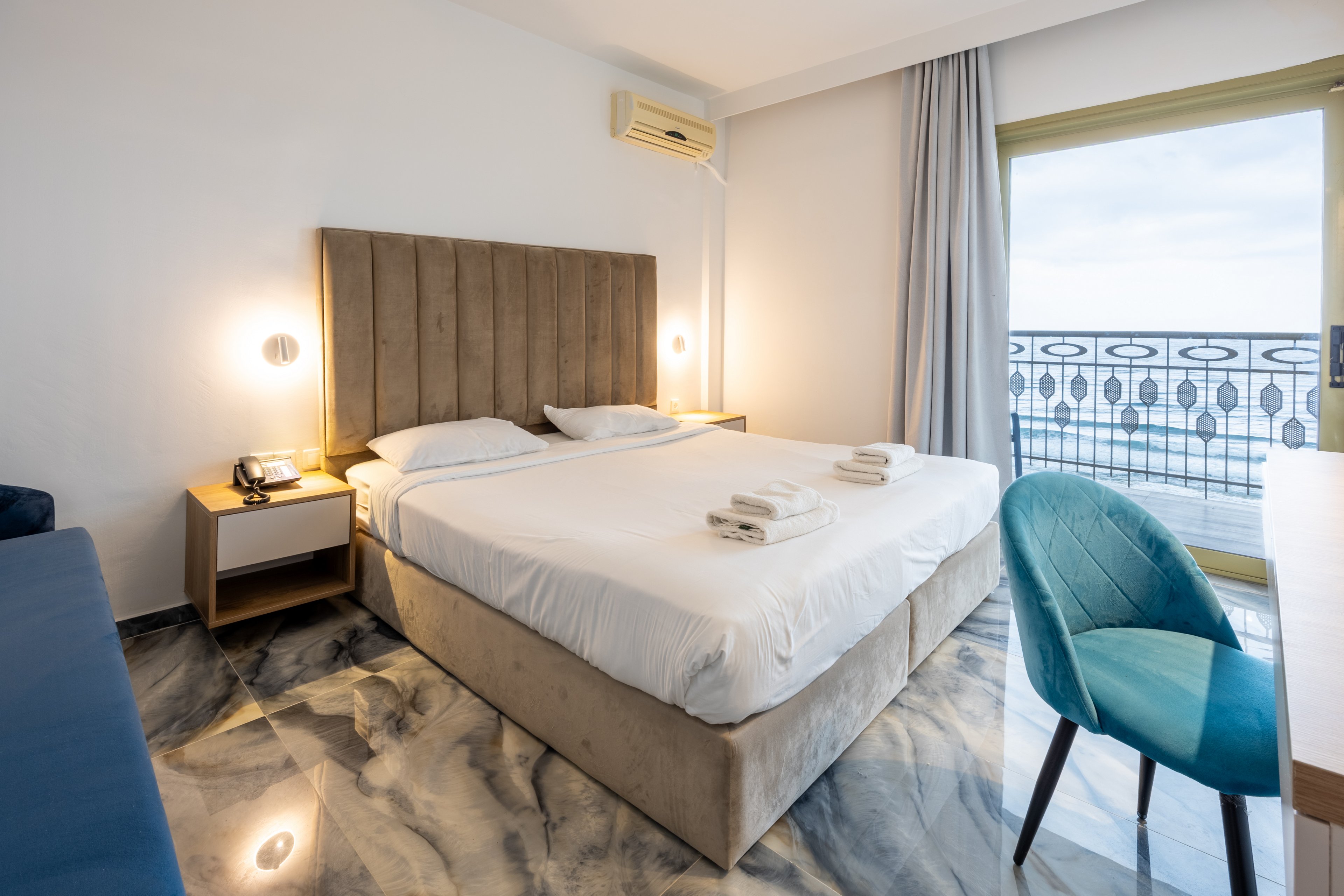 A contemporary hotel bedroom features a large double bed with a tall upholstered headboard and neatly folded white towels on top. The room is decorated in neutral tones with marble-style flooring, soft lighting from bedside lamps, and a blue accent chair by a wooden desk. A sliding glass door opens to a balcony with ornate metal railings overlooking the sea and sky. Minimalist furniture and clean lines create a calm, inviting atmosphere ideal for relaxation.