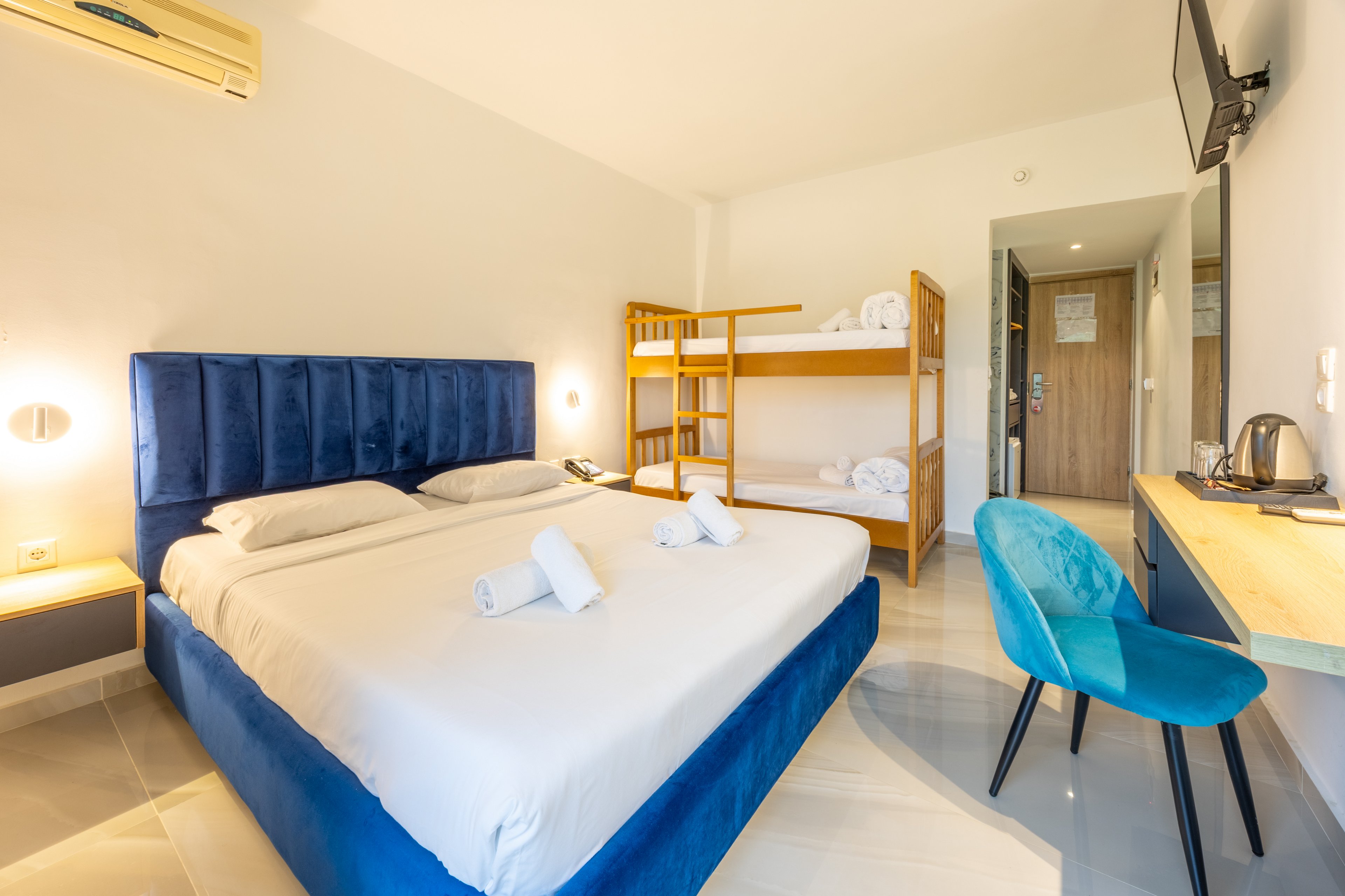 Bright, modern hotel room featuring a large double bed with a blue upholstered headboard and neatly rolled white towels. A wooden bunk bed stands against the wall, prepared with fresh linens for additional guests. The room includes a small work desk with a blue chair, a wall-mounted television, and a kettle station. Light-colored floors and walls, along with warm lighting, create a clean and welcoming atmosphere.