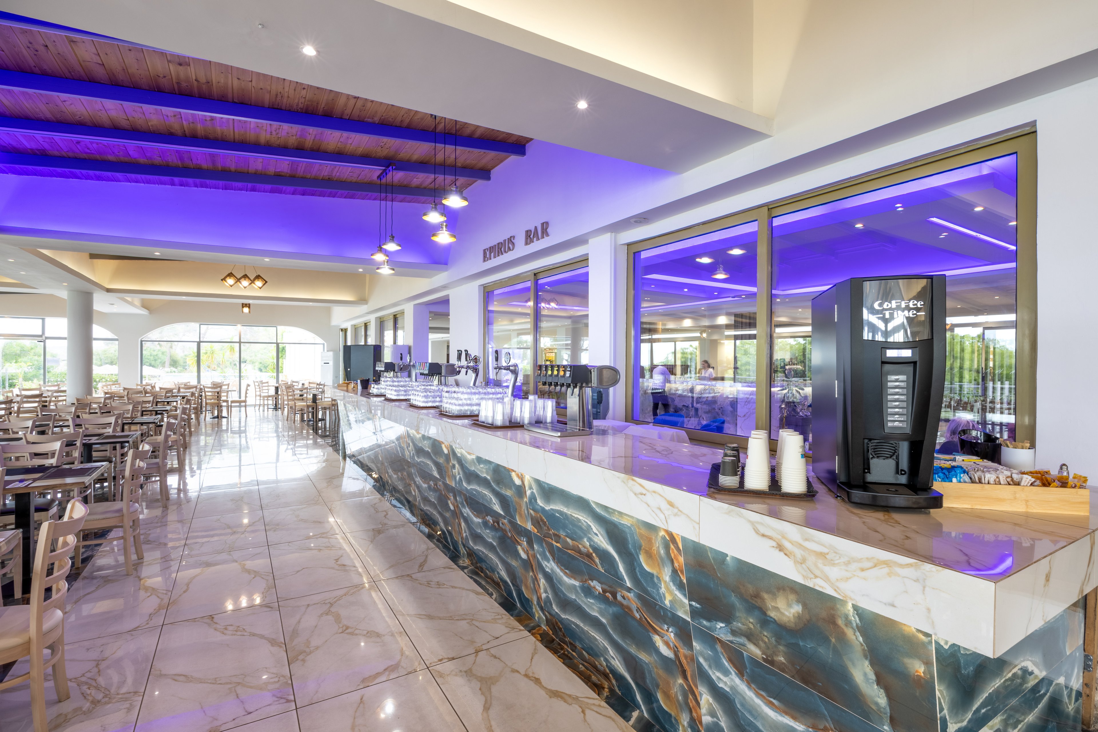 Spacious hotel dining room features a long marble counter labeled as a drinks bar. A self-service beverage machine and rows of cups are arranged neatly along the bar. Numerous empty tables and chairs fill the bright interior, illuminated by ceiling lights and purple accent lighting. Large windows and glass doors at the far end open to a green outdoor view, creating an inviting atmosphere.