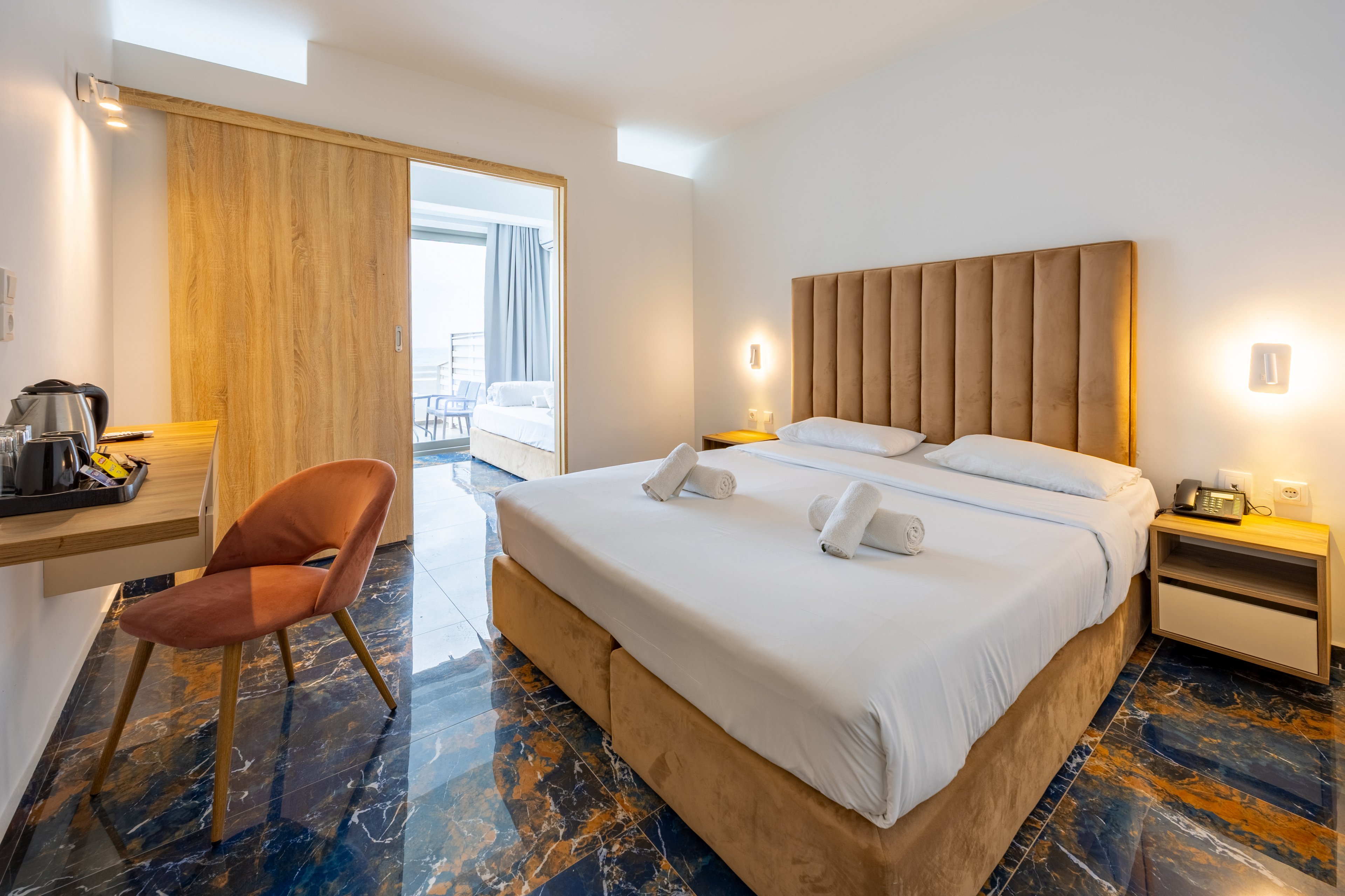 A contemporary hotel room features a large double bed with a tall upholstered headboard and neatly folded white towels. The space includes a wooden desk with a kettle and cups, a cushioned chair, and matching bedside tables with wall-mounted lights. Glossy marble-style flooring adds a luxurious touch to the minimalist white walls and warm wood accents. A sliding wooden door opens to an adjoining room or balcony area with natural daylight streaming in.