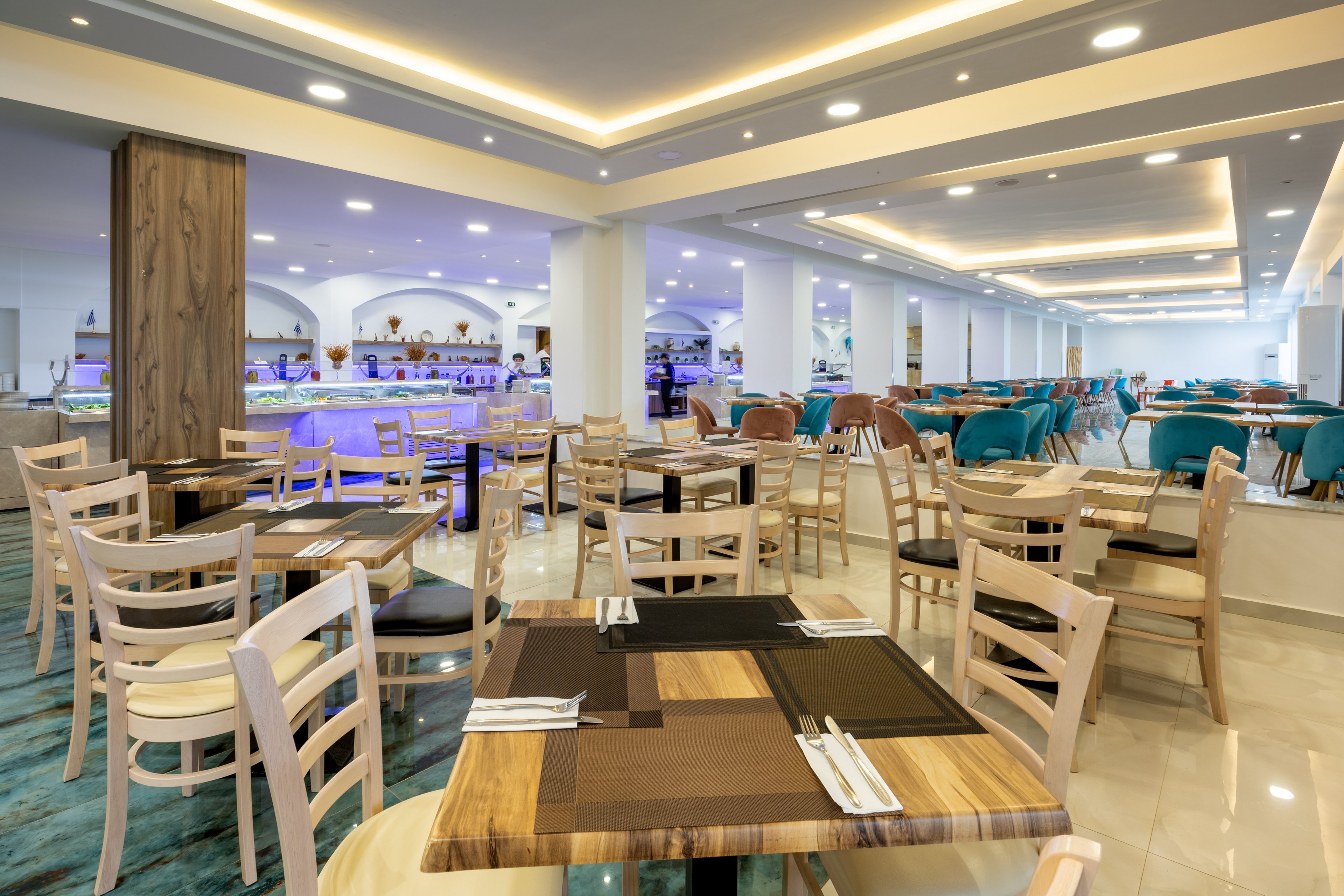 A spacious contemporary restaurant interior features neatly arranged wooden tables set with cutlery and placemats. Light wooden chairs surround the tables, and turquoise upholstered seating adds color to the space. Recessed ceiling lighting and glossy floors create a bright, clean atmosphere. In the background, a bar area with illuminated shelving and decorative arches enhances the modern hotel dining ambiance.