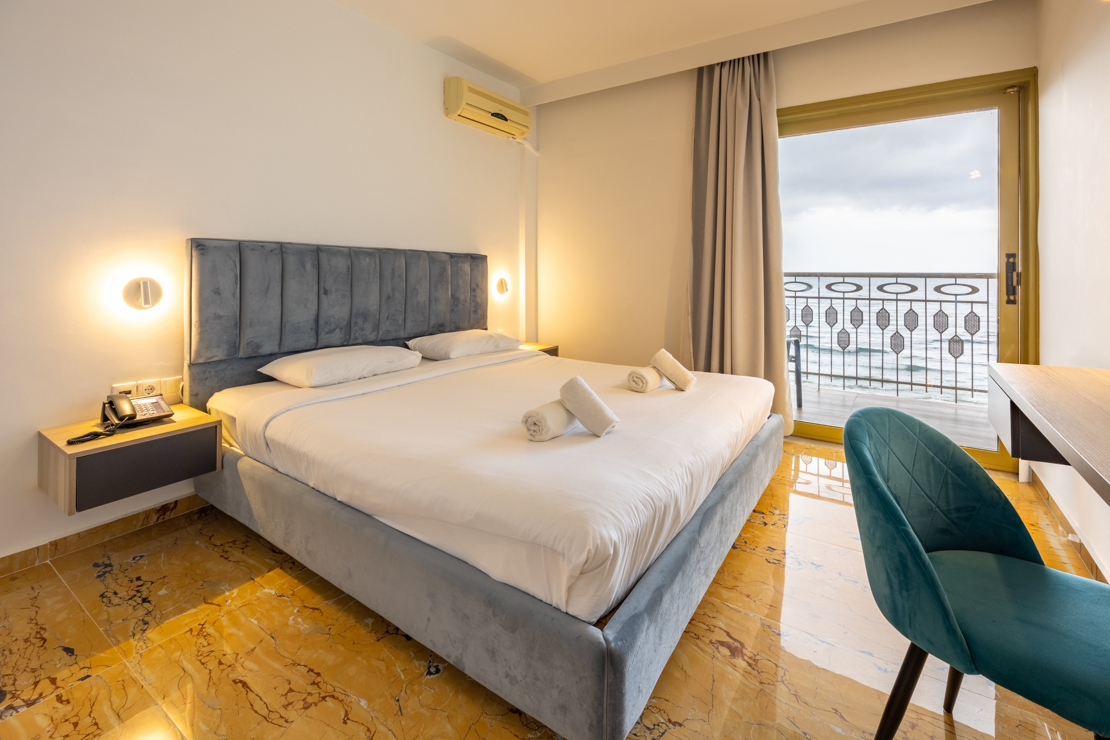 A contemporary hotel bedroom features a large neatly made bed with a soft upholstered headboard and rolled towels placed on top. Warm wall lights and a bedside table with a telephone add to the cozy, functional atmosphere. A teal chair sits by a wooden desk, while floor-to-ceiling curtains frame a glass door leading to a balcony with metal railings and an ocean view. The polished marble floor reflects the soft lighting, enhancing the clean and inviting feel of the room.
