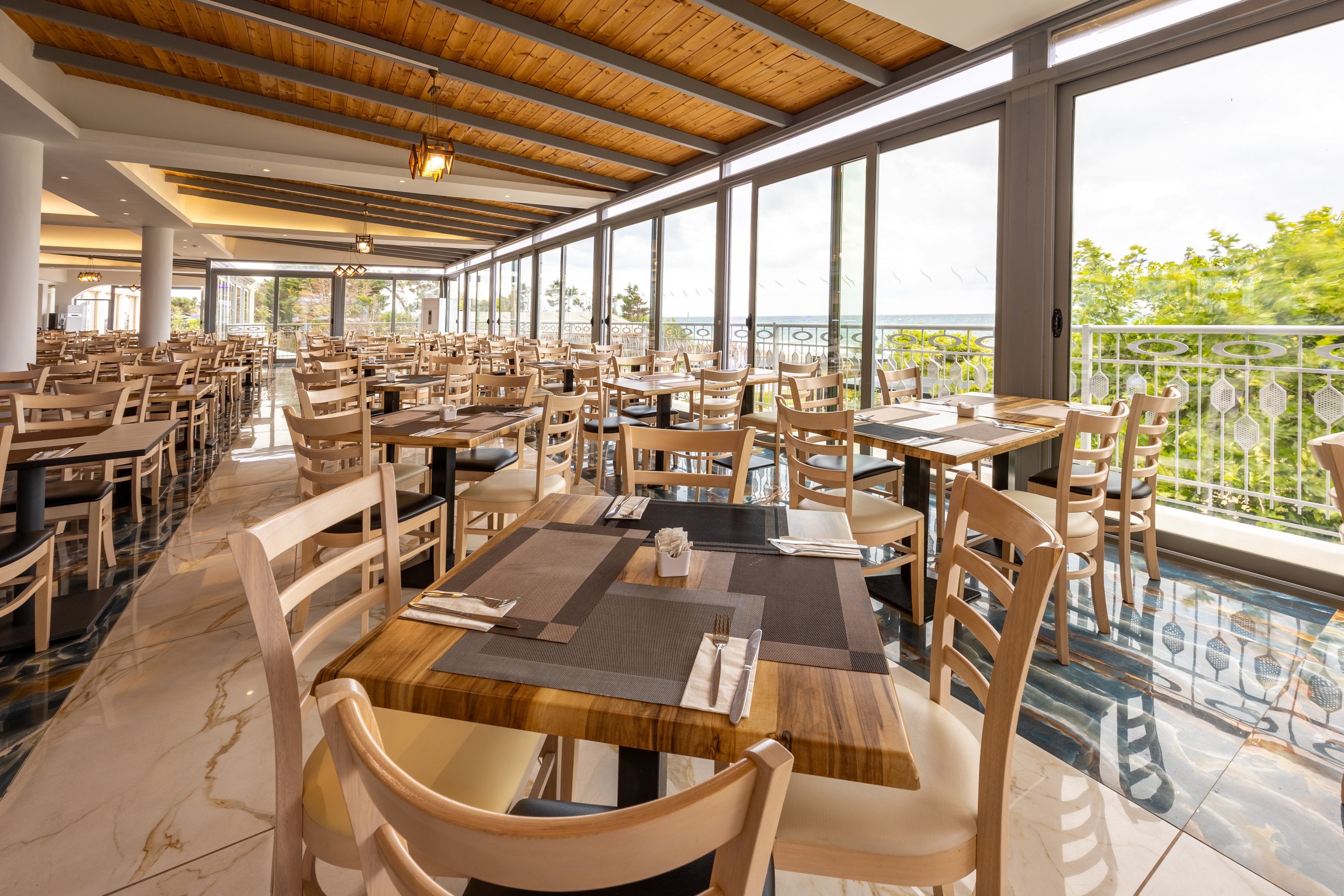 Spacious restaurant dining area with neatly arranged wooden tables and chairs is shown in warm natural light. Large floor-to-ceiling windows line one side, offering views of greenery and the distant sea. Place settings with cutlery and napkins are prepared on dark placemats, suggesting readiness for guests. A wooden ceiling and glossy tiled floor add to the airy, contemporary atmosphere.