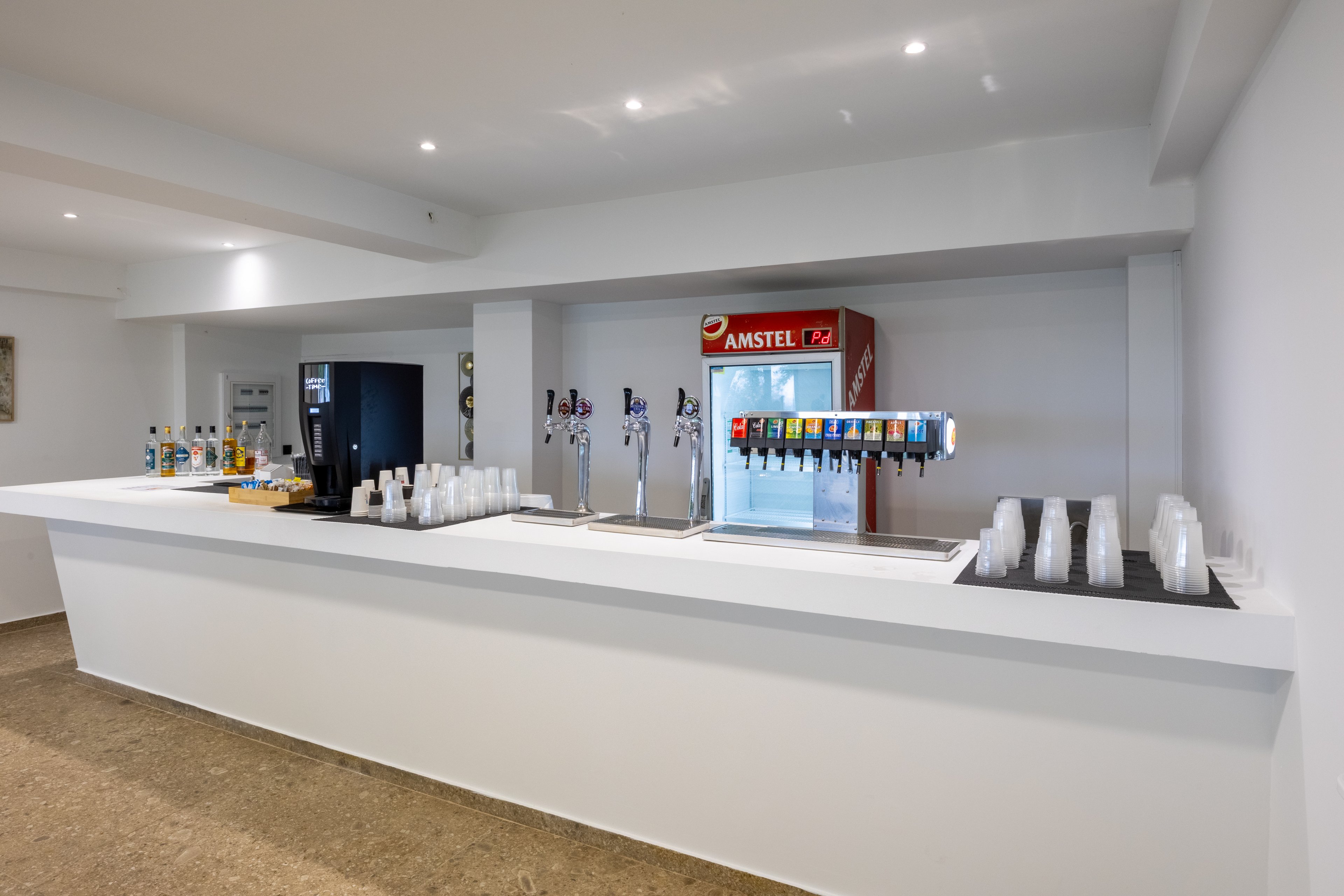 A clean, modern beverage bar features multiple drink dispensers and a long white counter. Soft drink fountain taps and beer taps are arranged centrally, with an Amstel-branded cooler visible in the background. Clear plastic cups are neatly stacked on both sides of the counter, ready for use. The bright, minimalist interior emphasizes functionality and a casual refreshment setting.