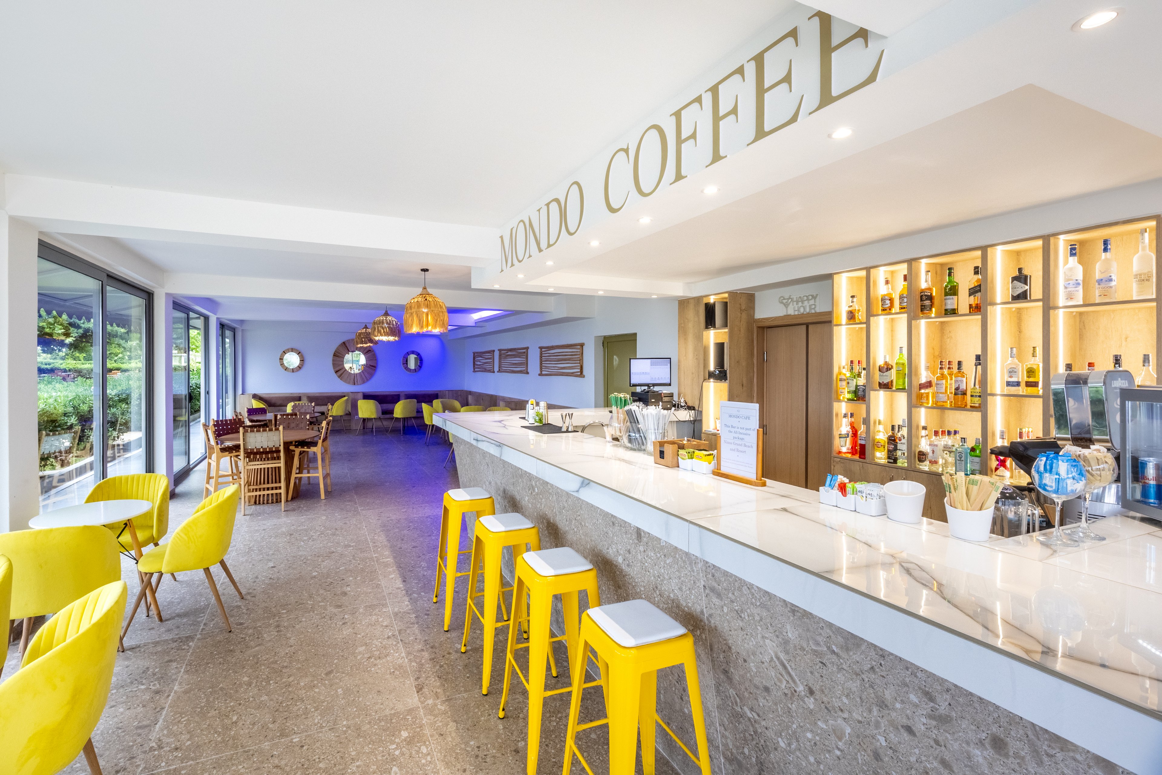 A contemporary coffee bar named Mondo Coffee features a long white marble counter with yellow bar stools. The interior is bright and airy, with large windows opening to greenery outside. Shelves behind the bar display various bottles and glassware, while a seating area with wooden tables and chairs extends into the background. Warm pendant lights and bold yellow chairs create a cheerful, inviting atmosphere.