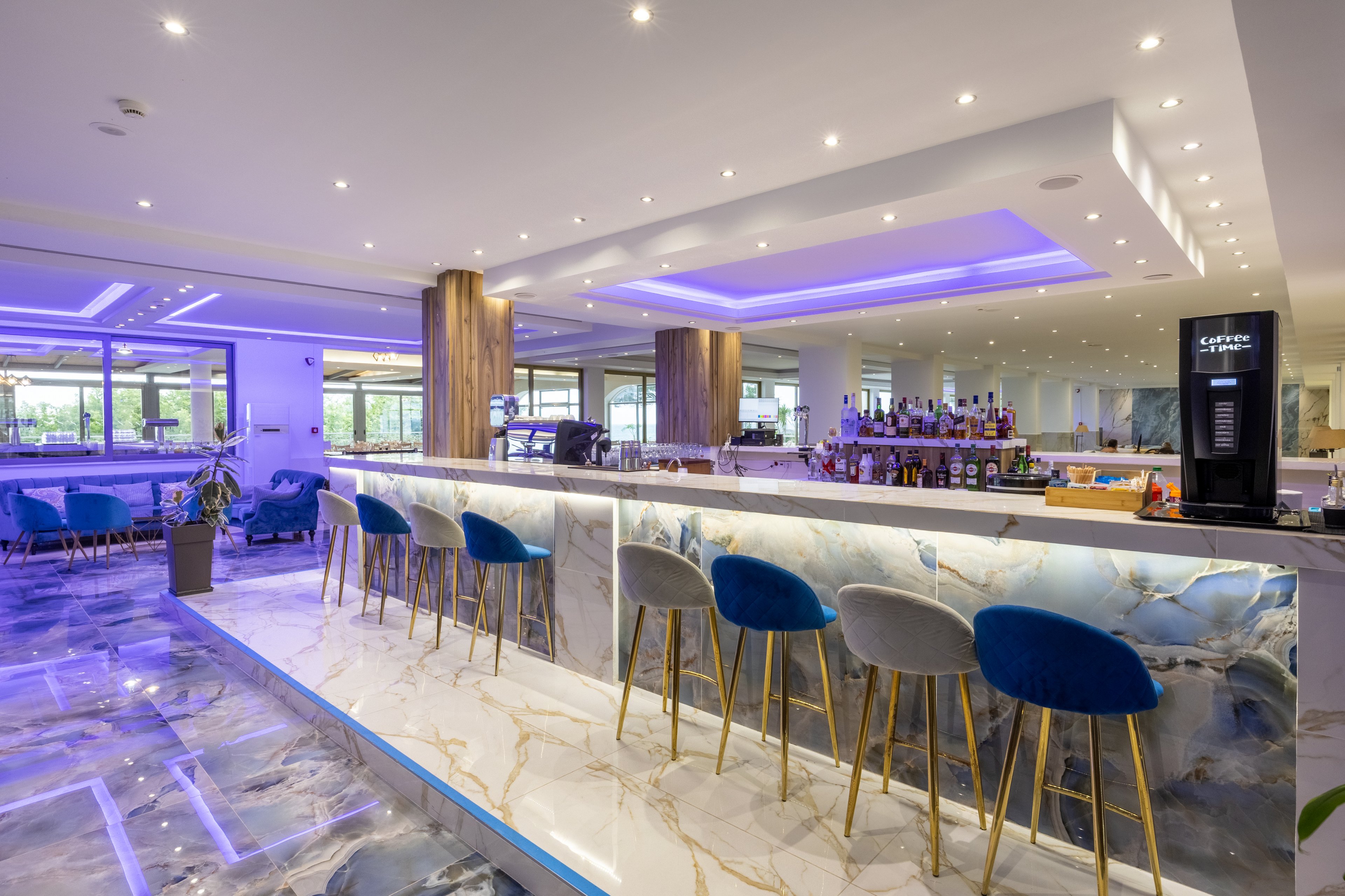 A contemporary hotel bar features a long marble counter with blue and gold veining and plush blue velvet bar stools. The space is brightly lit with recessed ceiling lights and purple LED accent lighting that reflects off the glossy floor. Bottles and glassware are neatly arranged behind the bar, with large windows providing views of greenery outside. The overall atmosphere is sleek, upscale, and inviting for evening drinks or social gatherings.