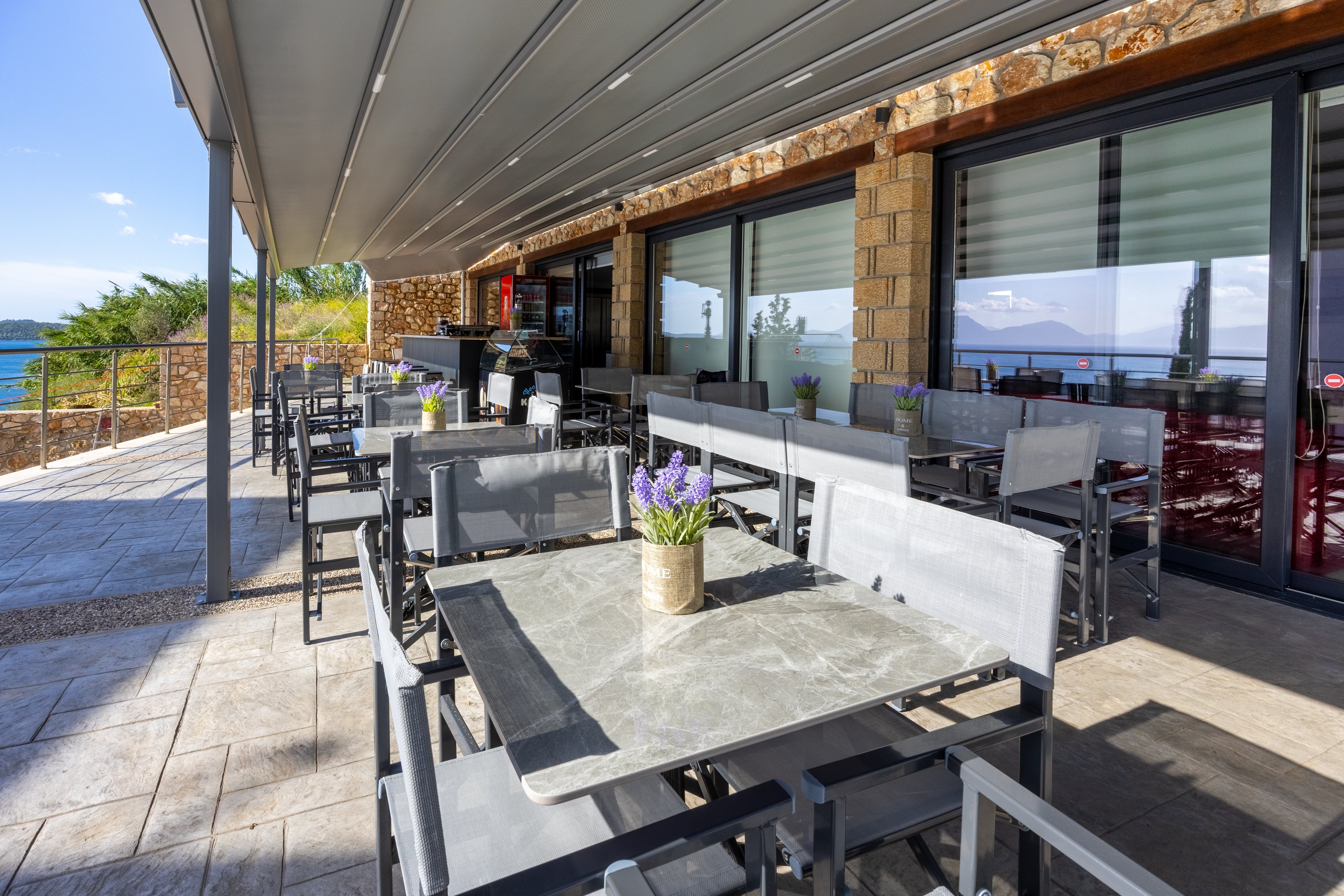 An outdoor restaurant terrace features empty tables and chairs under a modern metal awning. A small potted lavender arrangement decorates the foreground table, adding a touch of color. Large glass doors reflect the bright sky and surrounding landscape, while stone walls give the building a rustic feel. In the background, the terrace overlooks a scenic coastal view with blue water and distant hills.