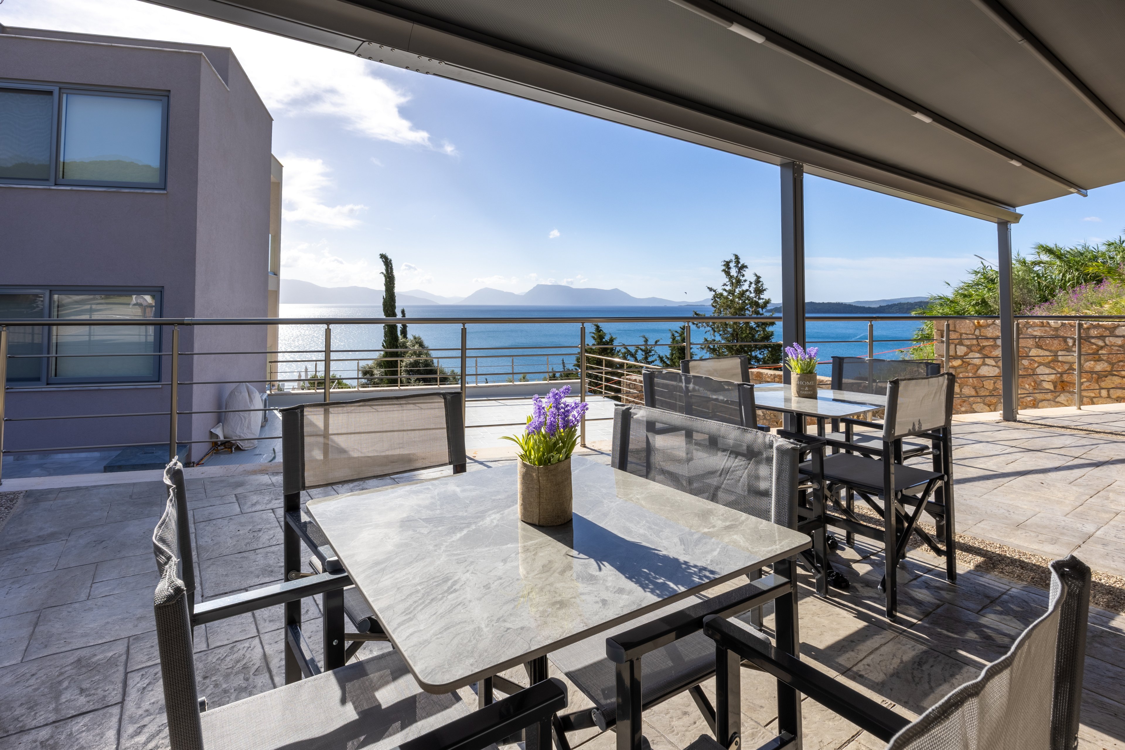 A modern outdoor terrace features metal and stone tables with black chairs arranged for al fresco dining. Potted purple flowers decorate the tabletops, adding a touch of color to the neutral surfaces. A large awning provides shade while railings frame a clear view of the blue sea and distant mountains. The bright daylight and calm water create a relaxed, vacation-like atmosphere.