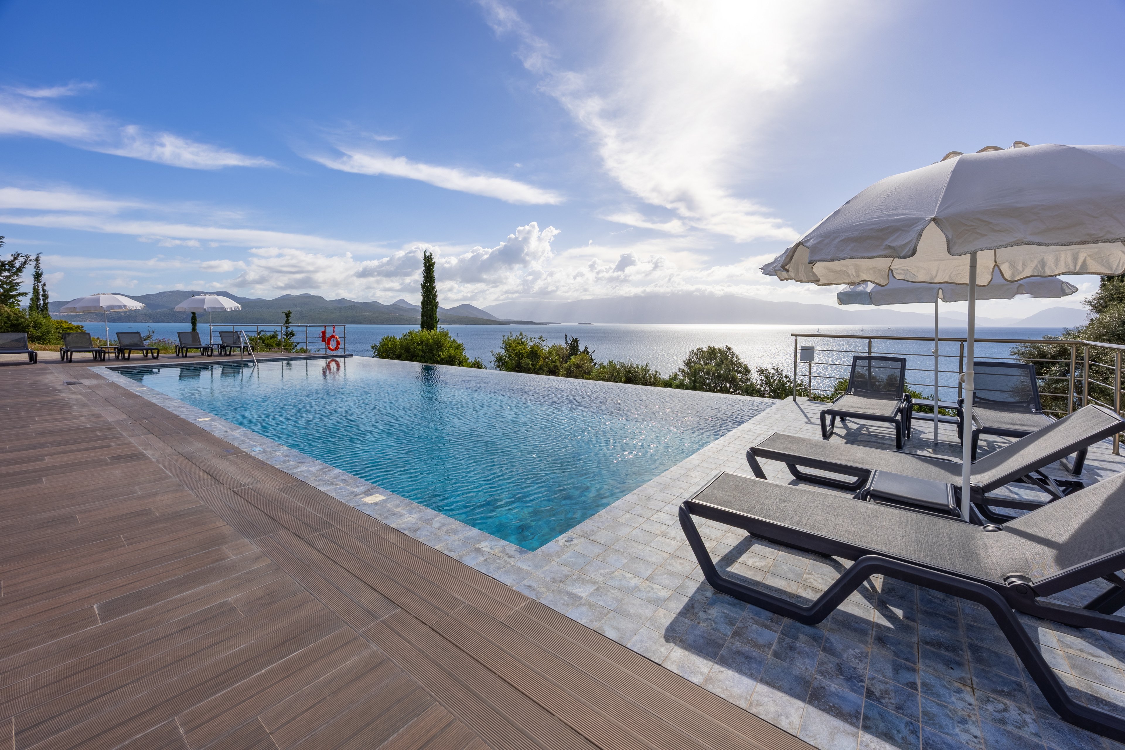 An elevated infinity pool stretches toward a calm, expansive sea under a bright sky. Sun loungers and large white umbrellas line the tiled poolside terrace, suggesting a relaxing resort setting. Distant hills and scattered clouds create a serene coastal backdrop. The overall scene conveys leisure, sunshine, and peaceful vacation vibes.