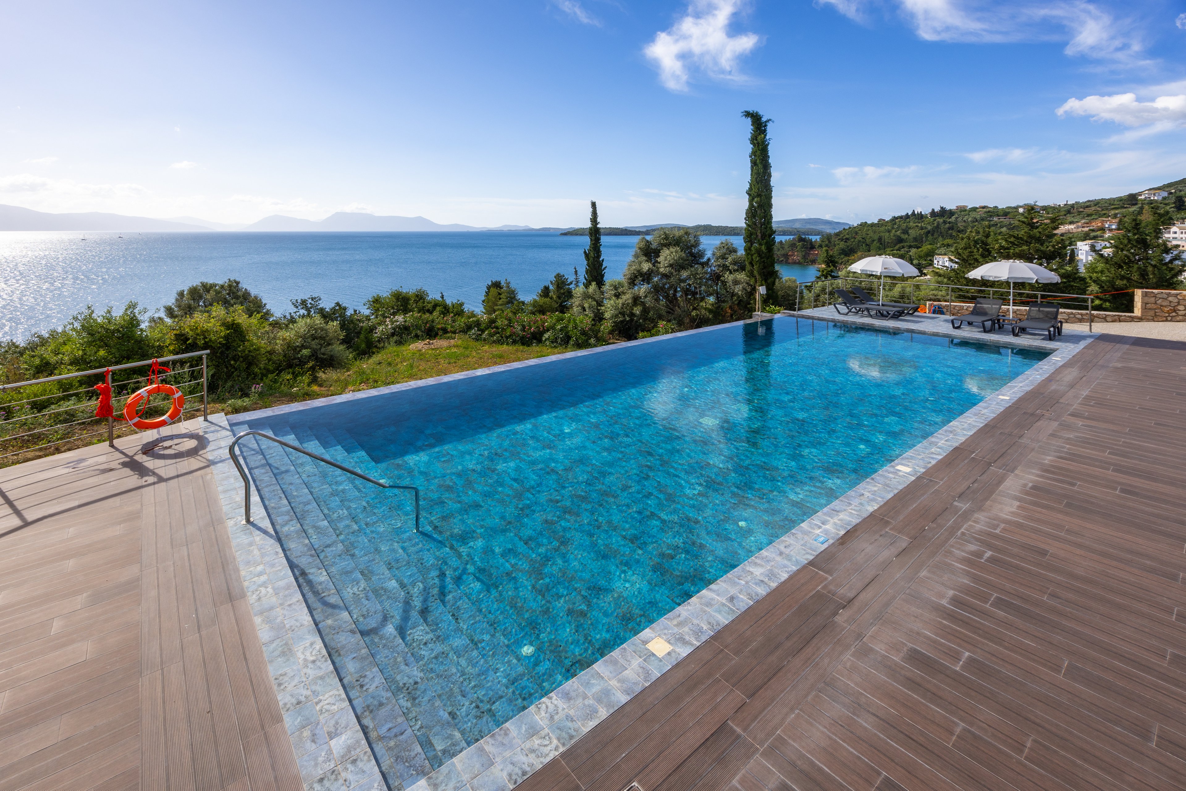 An infinity pool stretches toward a scenic coastal horizon, blending seamlessly with the blue sea beyond. The pool is surrounded by a spacious wooden deck with metal railings and a visible life preserver. In the background, green trees and shrubs lead down to the shoreline, with distant mountains across the water. A few sun loungers and umbrellas sit on the far side of the pool, suggesting a relaxing vacation setting under a clear, sunny sky.