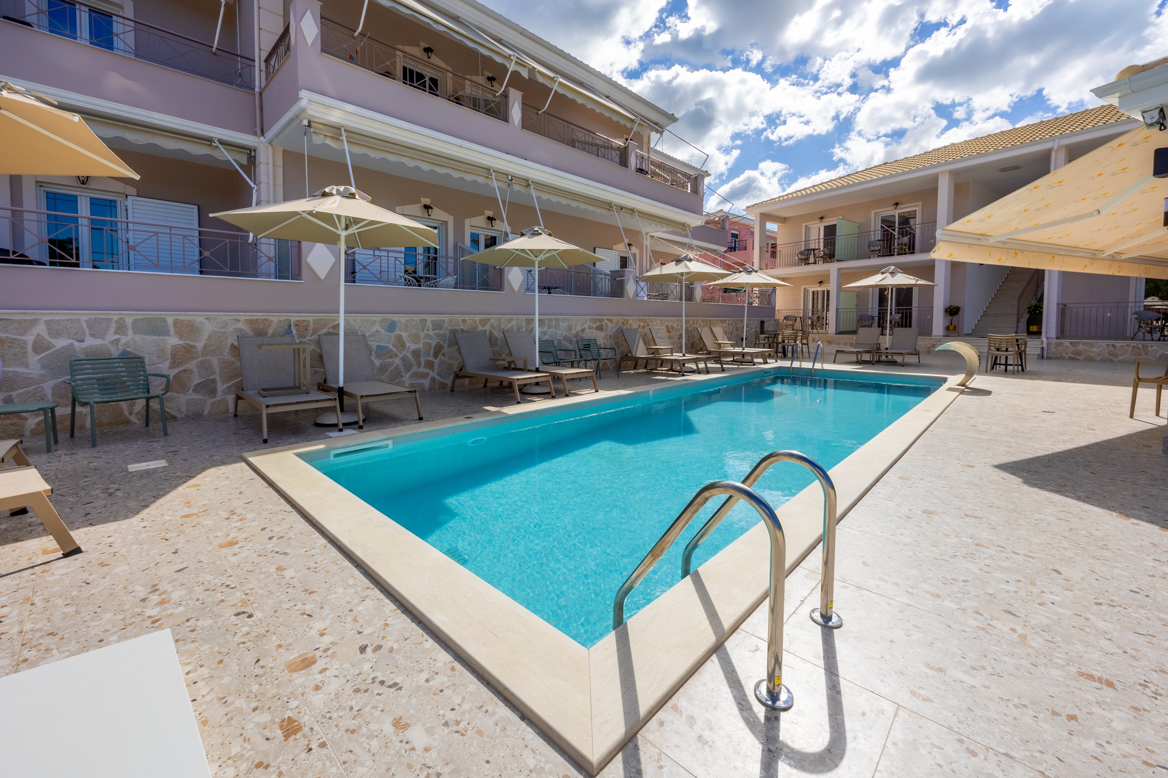 An outdoor swimming pool sits at the center of a small hotel courtyard. Lounge chairs and umbrellas line the poolside terrace, offering shaded seating areas for guests. Two-story buildings with balconies and stone accents surround the pool, creating an intimate resort atmosphere. Bright daylight and scattered clouds in the blue sky enhance the relaxed vacation mood.