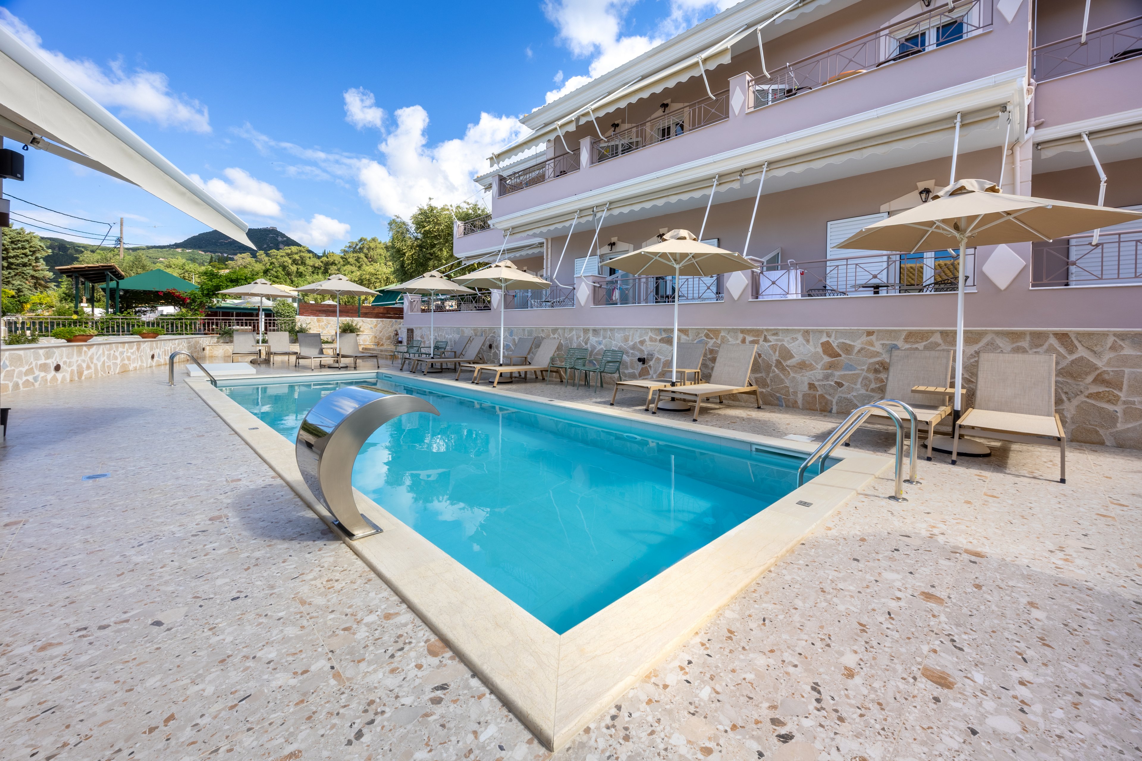 An outdoor swimming pool sits in the courtyard of a modern hotel or apartment building. Sun loungers and beige umbrellas line the poolside terrace, creating a relaxing vacation atmosphere. The water feature at the pool edge and stainless steel ladder add a contemporary touch. In the background, balconies, greenery, and distant hills are visible under a bright blue sky with scattered clouds.