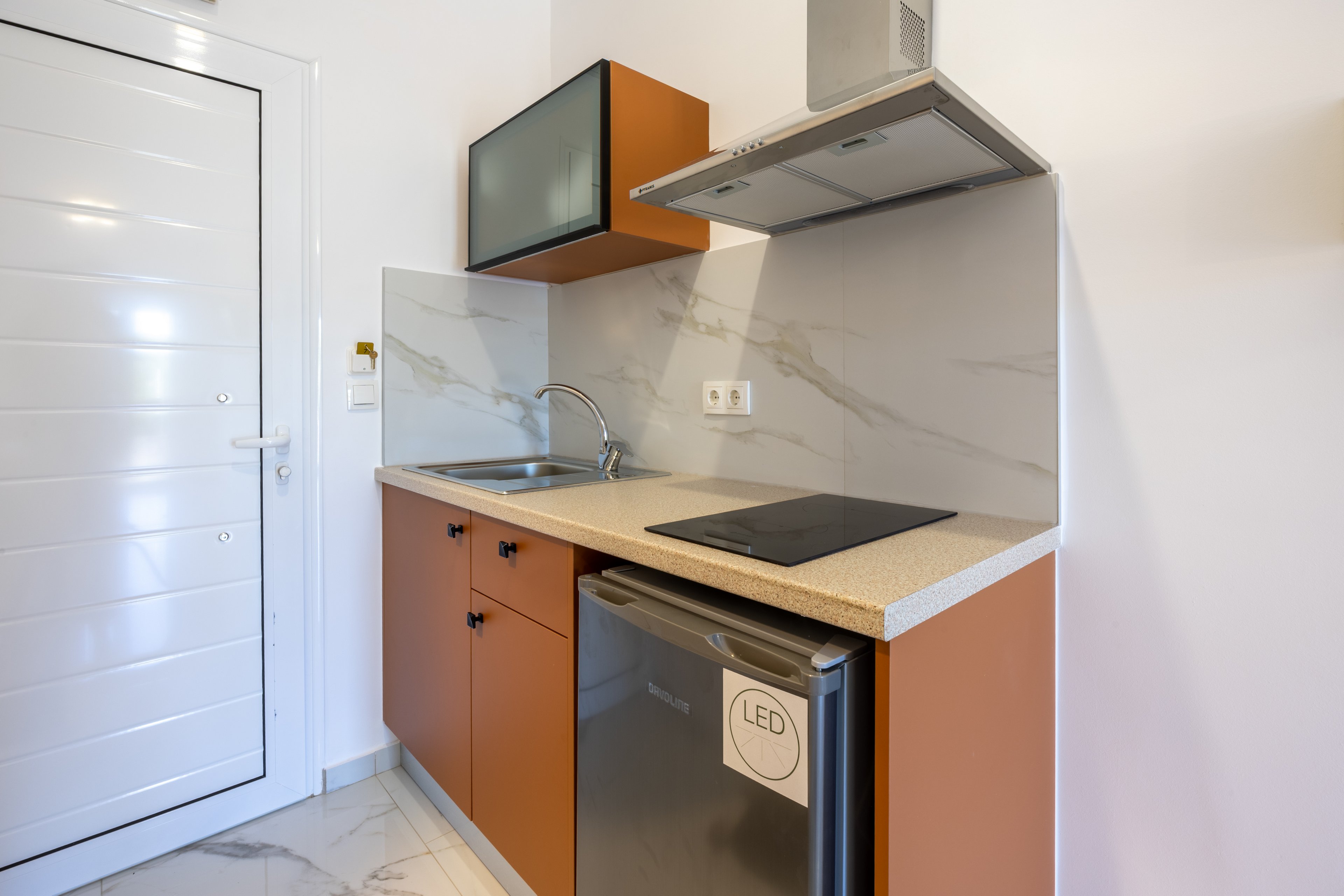 A compact modern kitchenette features brown cabinets, a small sink, and a sleek induction cooktop. A stainless steel mini fridge with an LED label sits beneath the countertop. Above the cooking area is a stainless steel range hood and an upper cabinet with a dark glass door. The space is finished with light marble-effect walls and flooring, creating a clean and minimalist look.