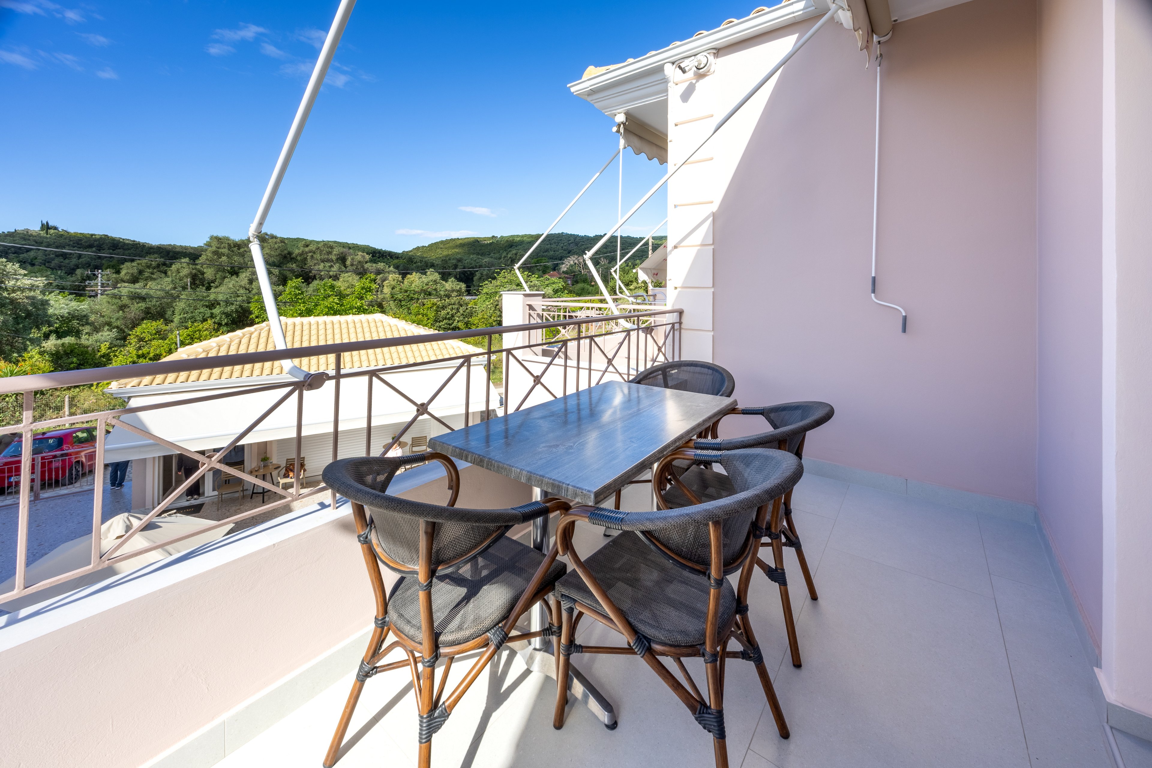 A bright balcony terrace features a sleek metal dining table surrounded by four dark chairs. The space overlooks nearby rooftops and lush green hills under a clear blue sky. Simple railings frame the view, and the pale walls and floor enhance the airy, minimalist feel. The scene suggests a relaxing outdoor spot for meals or socializing in warm weather.