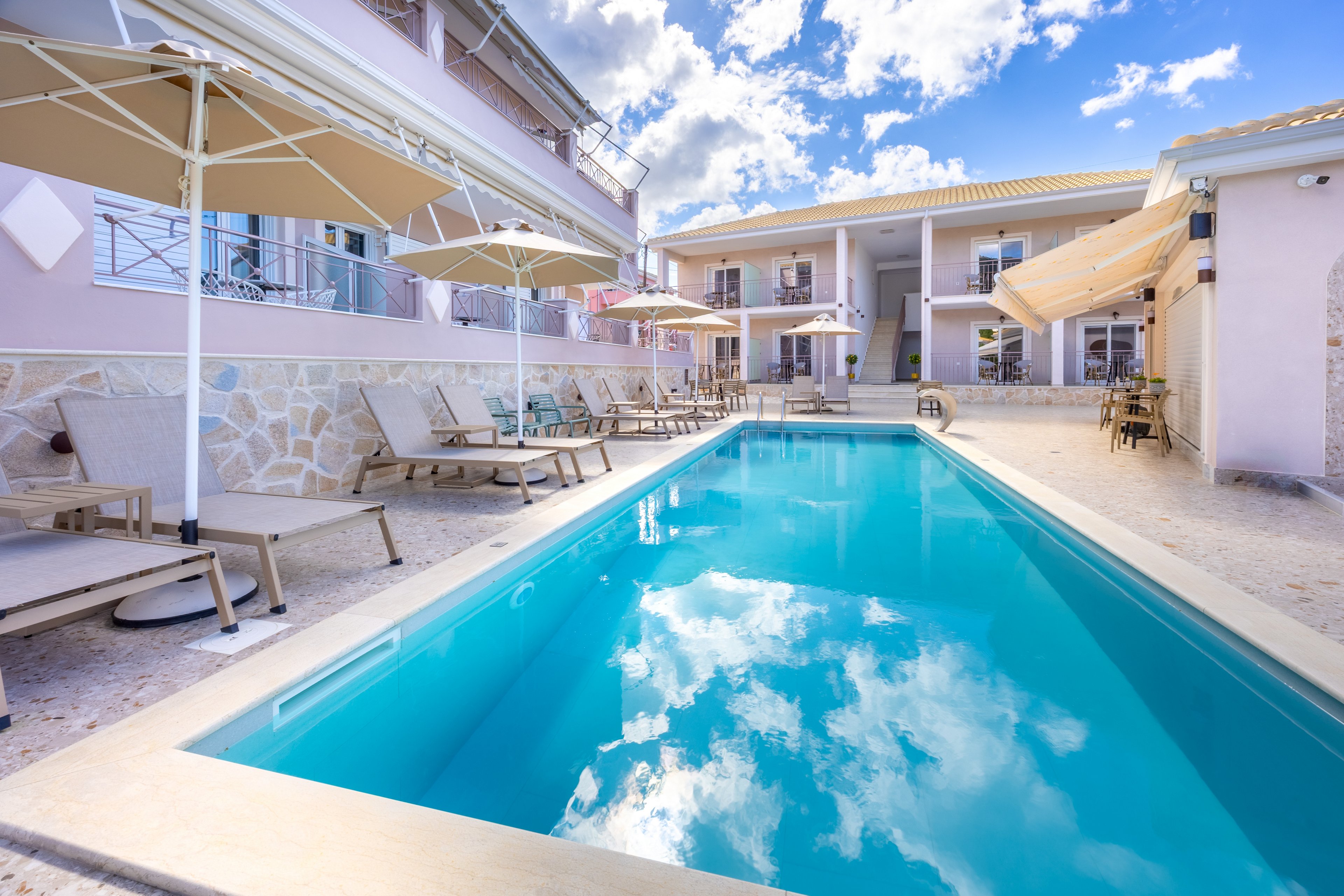 A bright outdoor swimming pool stretches between two light-colored hotel buildings under a partly cloudy sky. Sun loungers and large umbrellas line the poolside, creating a relaxing resort atmosphere. Reflections of the clouds are visible on the calm blue water, enhancing the serene mood. The scene suggests a peaceful vacation setting with comfortable seating and warm weather.