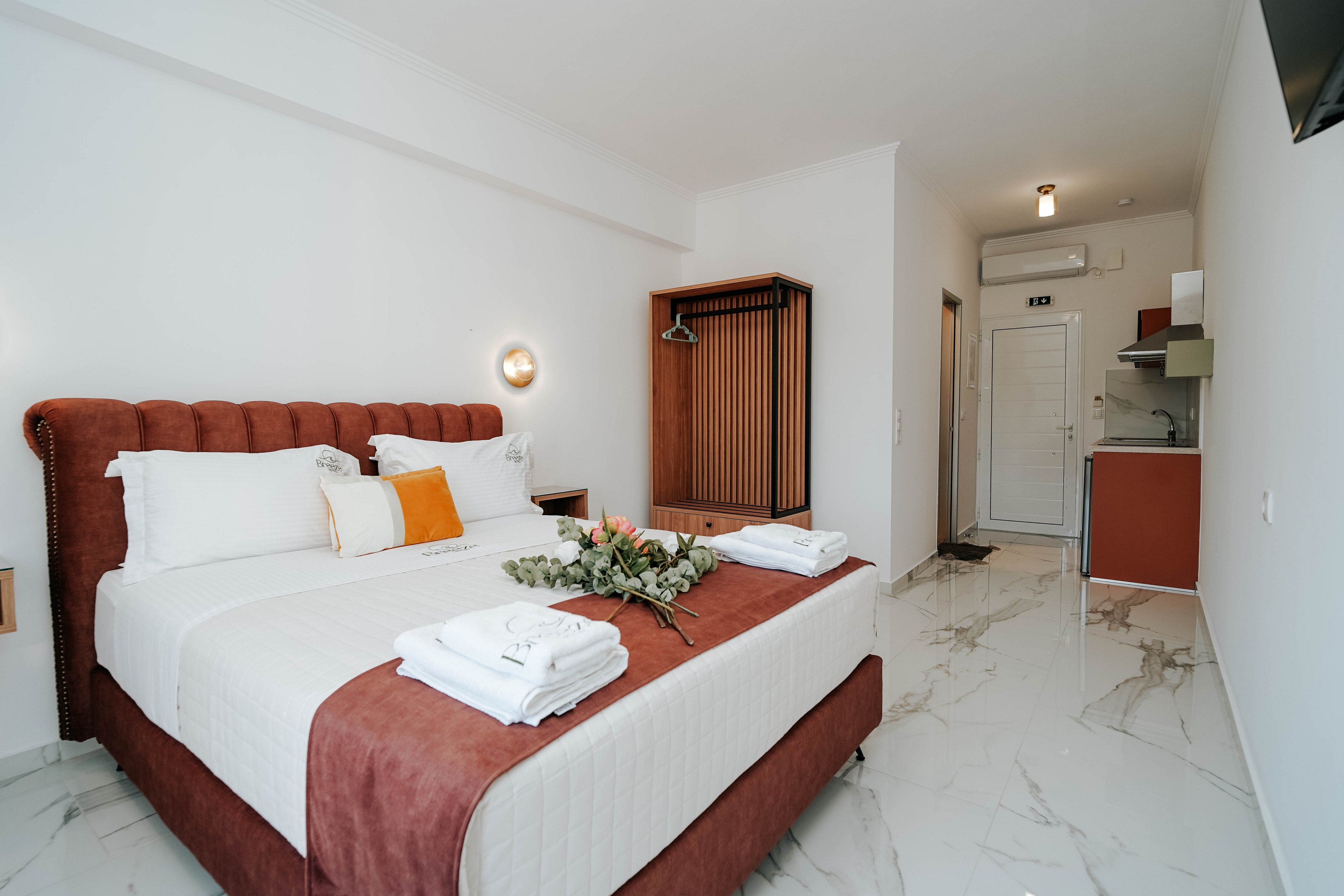A modern hotel room features a neatly made double bed with white linens and a rust-colored throw. Fresh towels and a small greenery arrangement are placed on the bed, adding a welcoming touch. The room has a bright, minimalist design with white walls, marble-look flooring, and warm wood accents. In the background, a compact kitchenette and wardrobe are visible near the entrance door, emphasizing functionality and comfort.