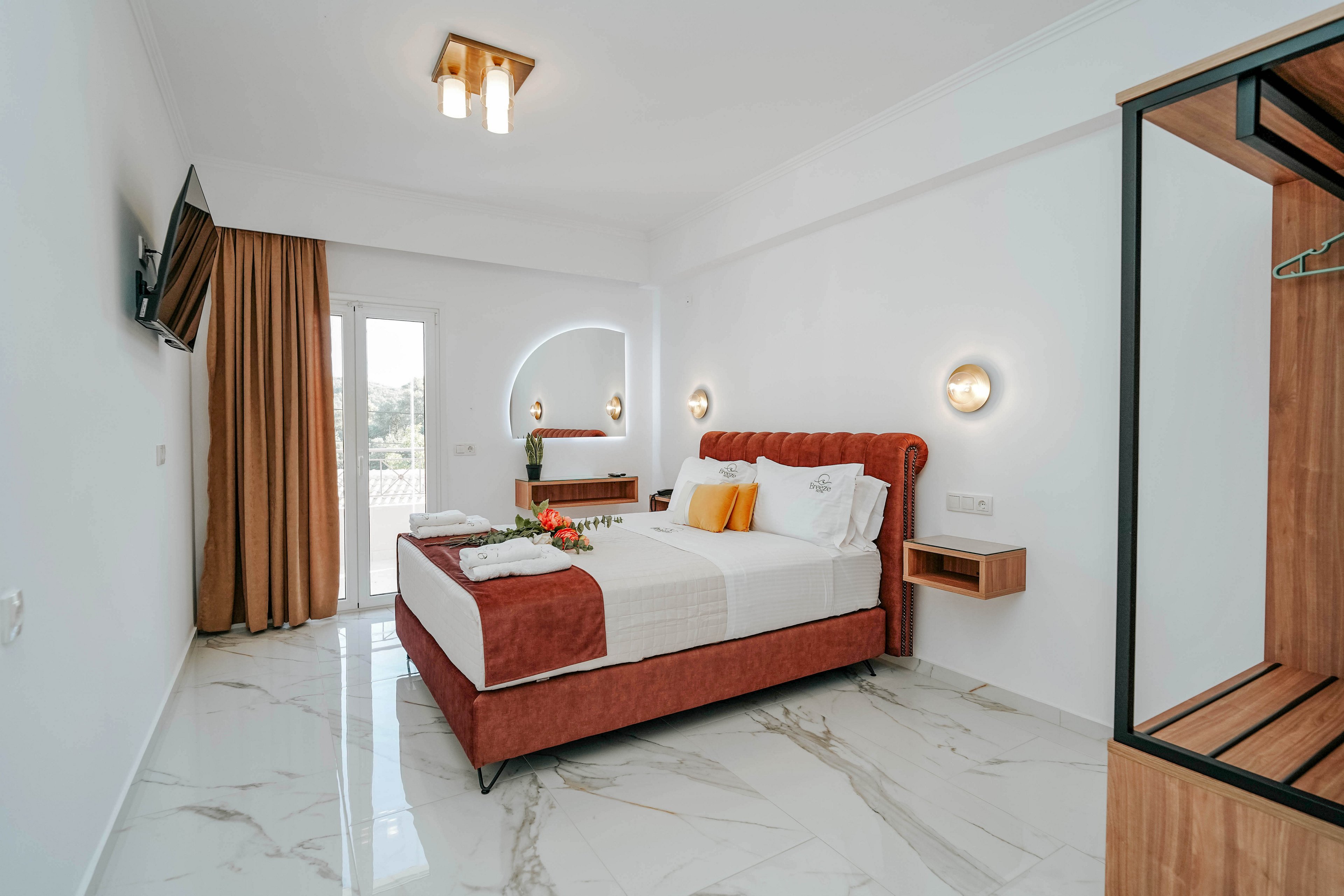 A contemporary hotel bedroom features a red upholstered bed with crisp white linens and accent pillows. The room is bright and minimalist, with white walls, marble-look flooring, and warm wood details. A wall-mounted television, bedside shelves, and a small open wardrobe add functional touches. Natural light enters through a glass door with brown curtains, creating an inviting and clean atmosphere.