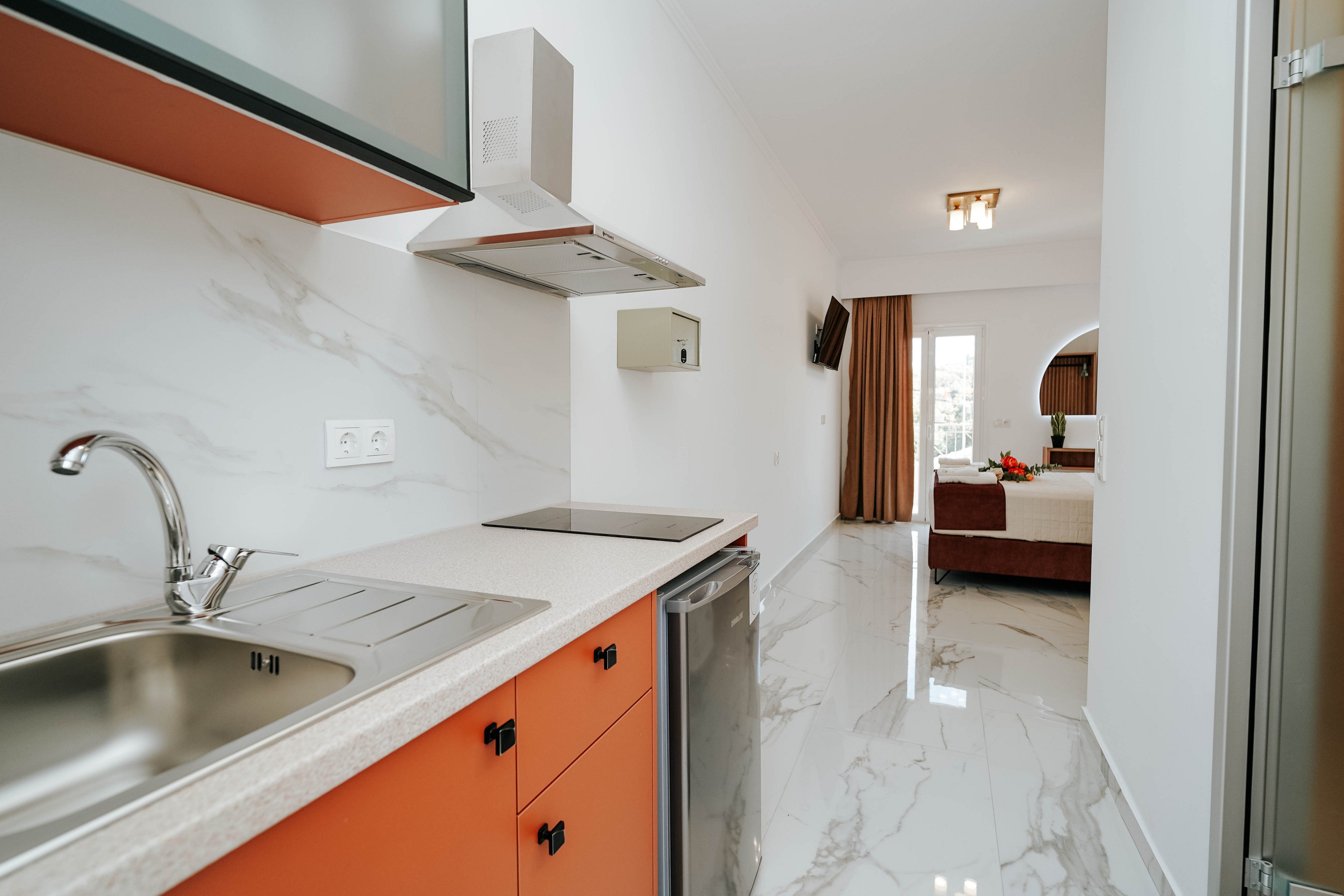A modern studio apartment features a compact kitchenette with orange lower cabinets, a stainless steel sink, and a small cooktop. The space has glossy white marble-style flooring that extends from the kitchen into the sleeping area. In the background, a neatly made bed, wall-mounted television, and large window with curtains create a cozy living space. The overall design is clean, minimal, and contemporary, emphasizing functionality in a small footprint.
