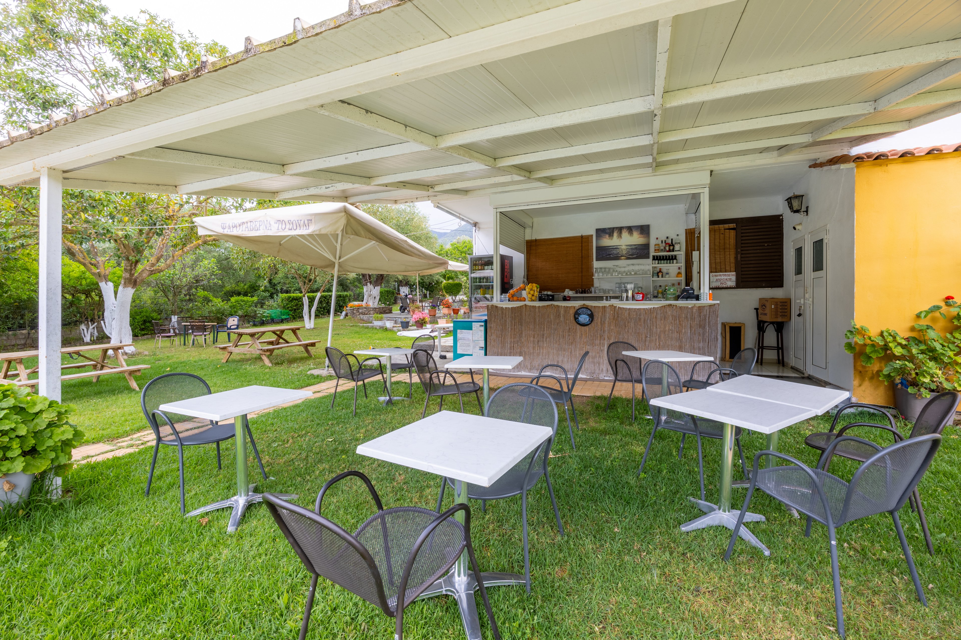 An open-air café features several empty metal tables and chairs arranged on a green lawn under a white pergola roof. The small bar counter in the background is stocked with drinks and snacks, with a staff area partially visible inside. In the garden beyond, large umbrellas shade wooden picnic tables and additional seating. Lush trees and plants surround the space, creating a relaxed and inviting atmosphere for outdoor dining.
