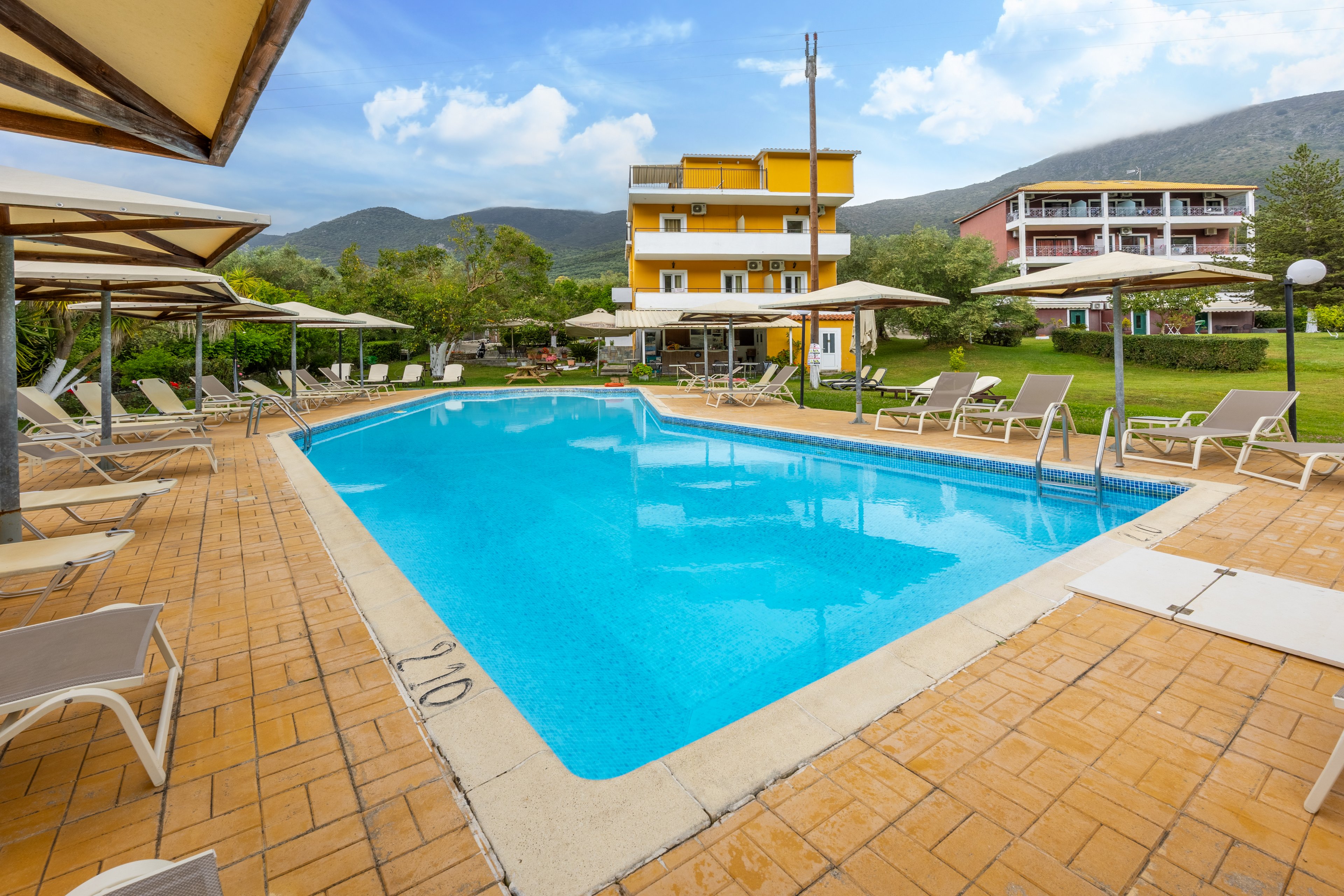 An inviting outdoor swimming pool is surrounded by beige sun loungers and parasols on a tiled terrace. A yellow multi-story hotel building stands at the far end of the pool, with another accommodation block visible to the right. Green lawns, trees, and distant hills frame the relaxing resort setting under a bright, partly cloudy sky. Depth markings such as 2.10 are visible on the pool edge tiles.
