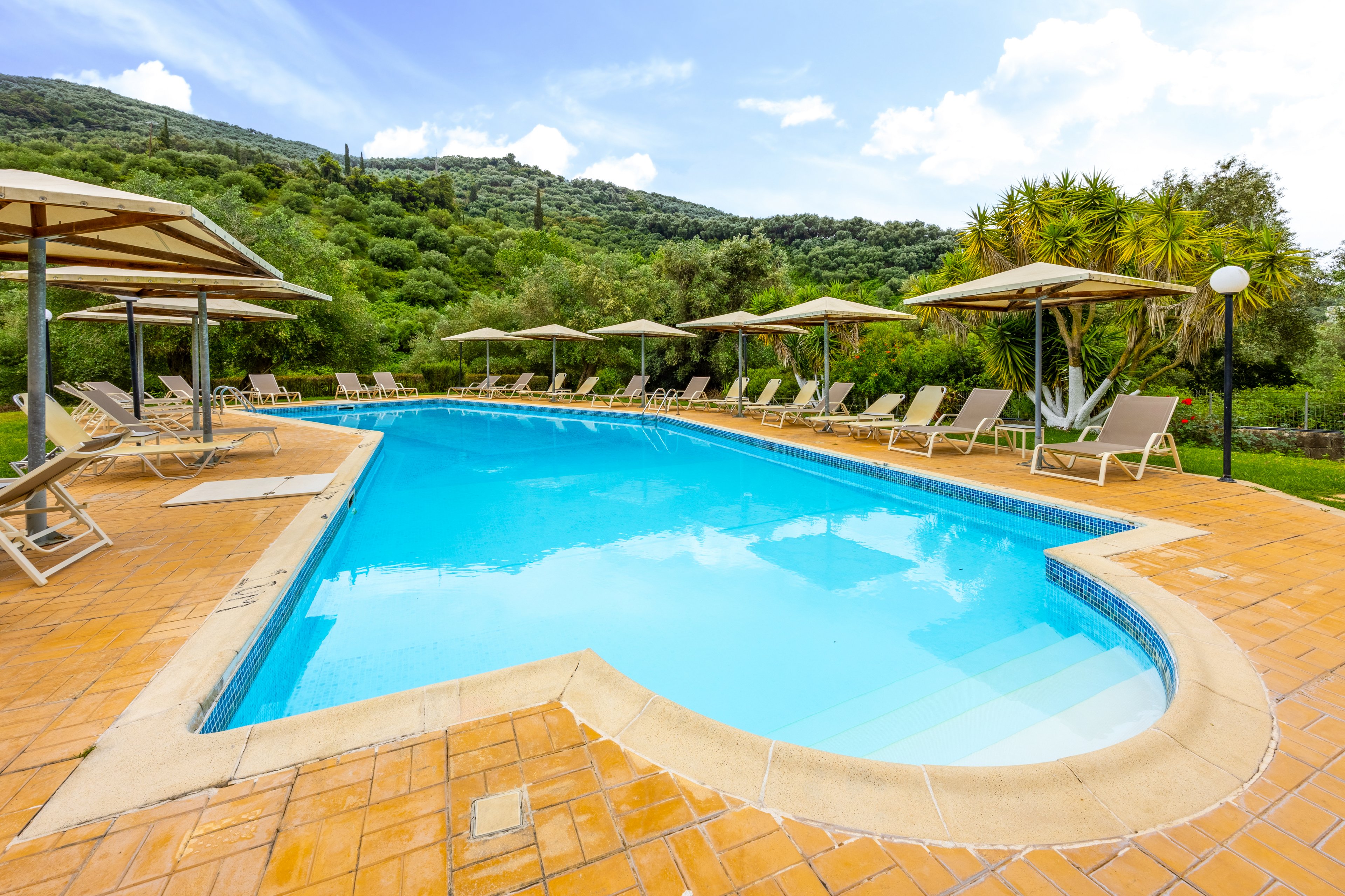 An inviting outdoor swimming pool is surrounded by tan lounge chairs and umbrellas on a tiled deck. The pool sits in a lush green landscape with trees and hills in the background. Clear blue water reflects the sky, creating a bright and refreshing atmosphere. The scene suggests a peaceful vacation setting at a resort or hotel.