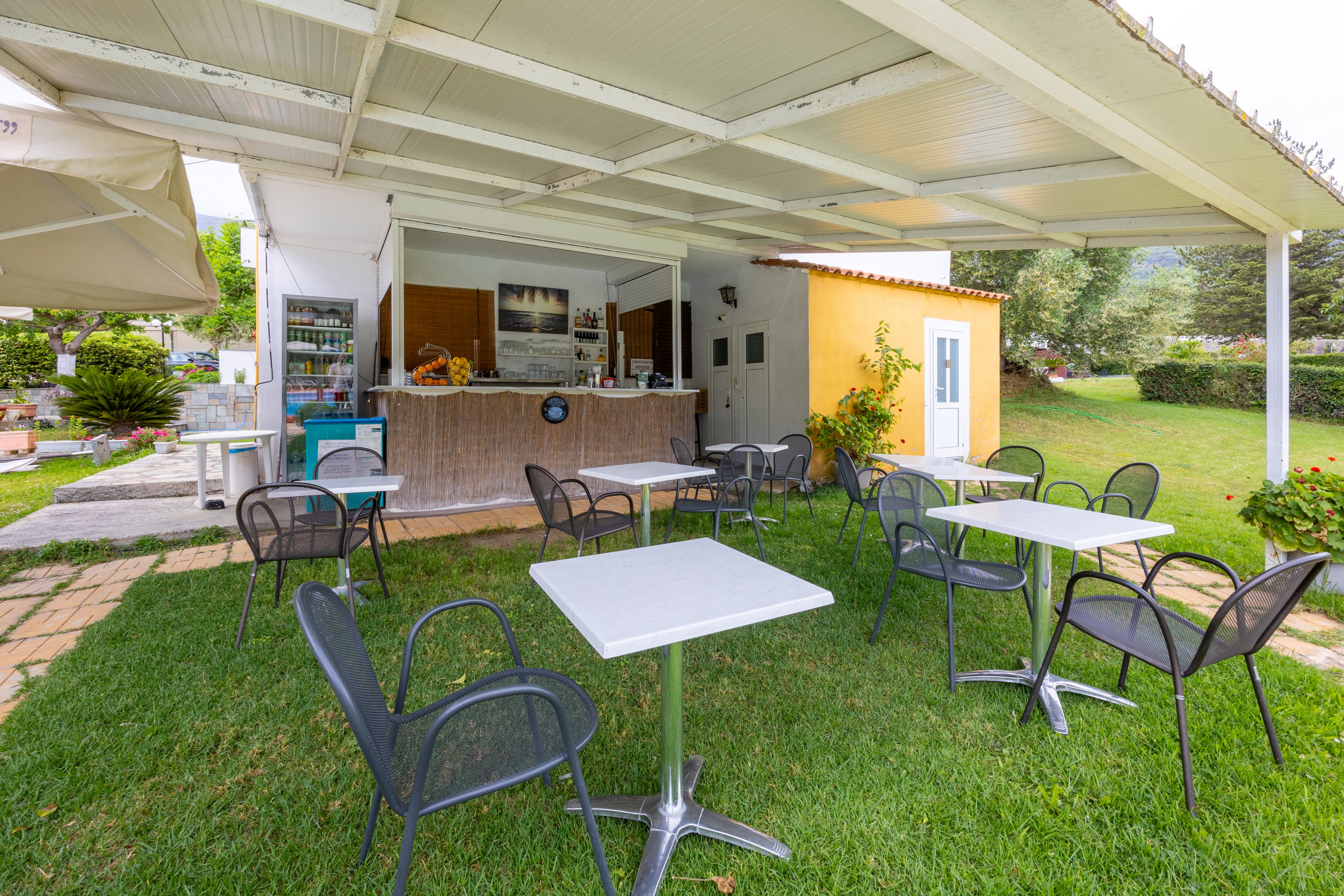 An open-air café features several empty metal tables and chairs arranged on a green lawn. The bar counter sits under a white roofed structure, with drinks and supplies visible behind it. A small yellow building with a white door stands to the side, adding contrast to the scene. Trees and shrubs surround the area, creating a relaxed, casual outdoor atmosphere.