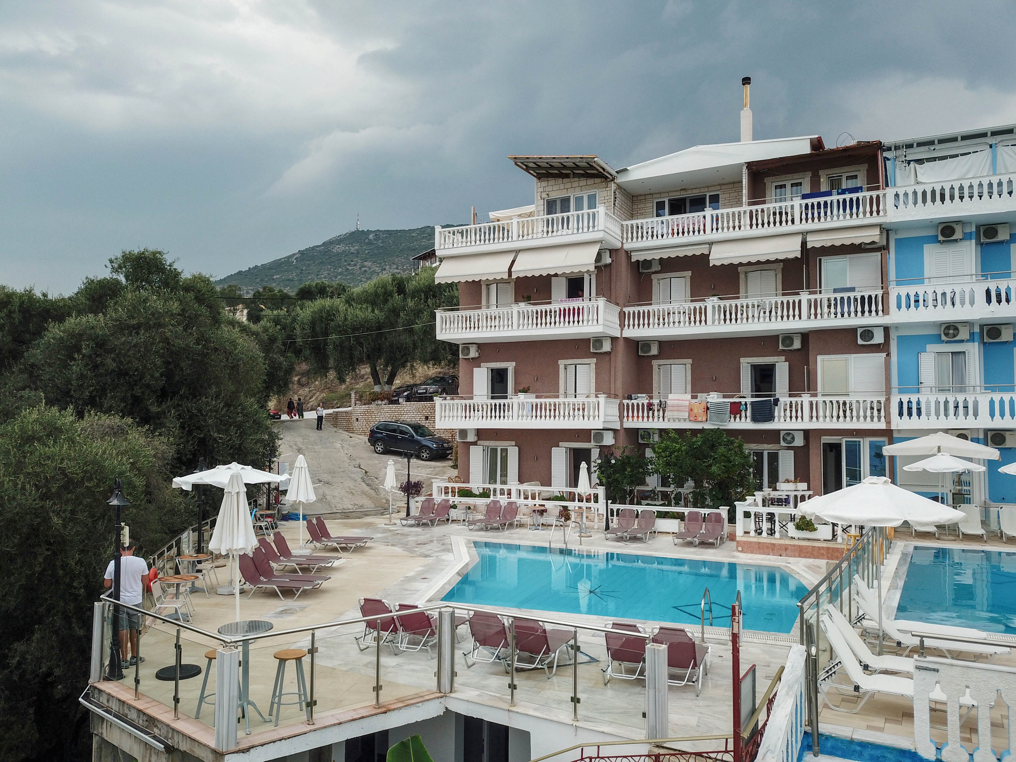 A multi-story resort hotel features balconies and a large outdoor swimming pool surrounded by lounge chairs and umbrellas. The setting is outdoors with a mountainous backdrop and overcast skies, suggesting an impending storm. The pool area is empty except for one person standing near the edge, and several parked cars are visible in the background. The building exterior is painted in pastel colors, adding a relaxed vacation atmosphere.