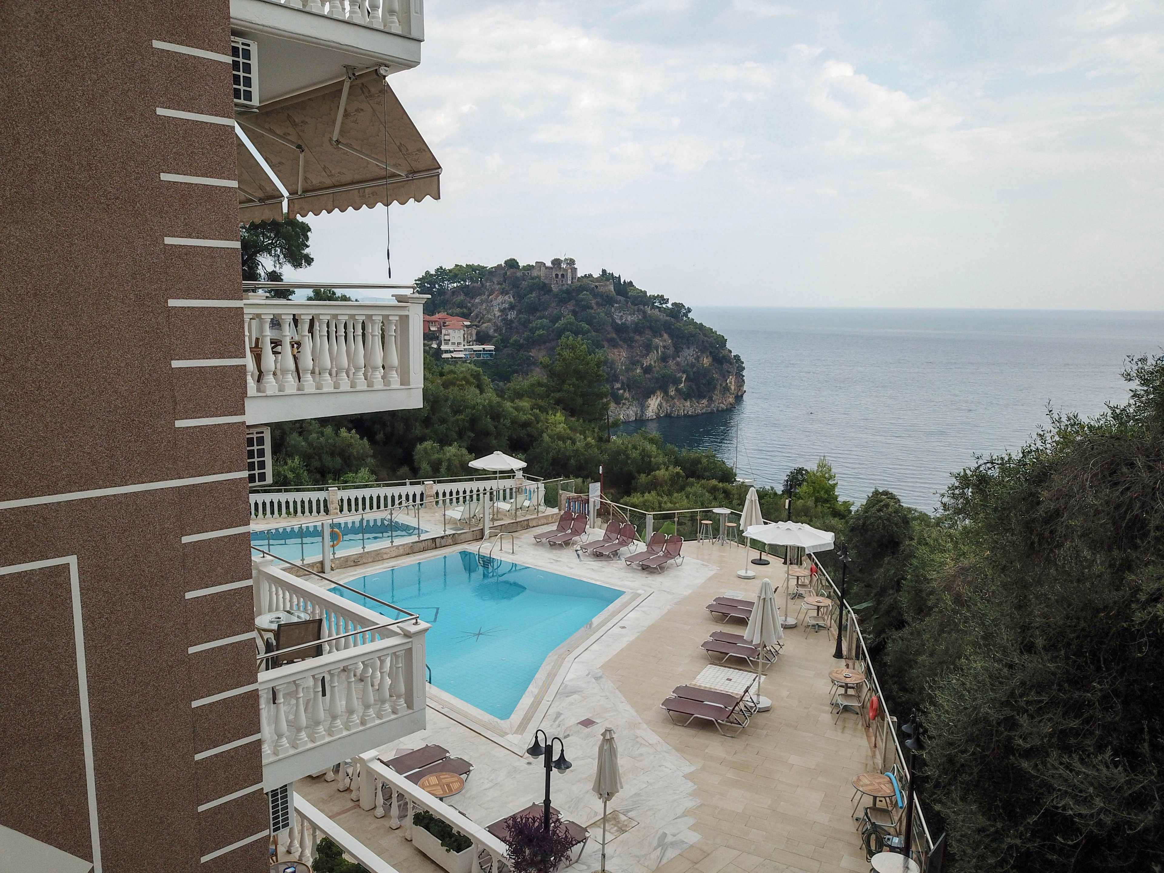 A hotel pool area overlooks a dramatic cliff and expansive sea, with several sun loungers and umbrellas arranged around the pool. The setting is tranquil, with lush greenery and a partially cloudy sky. The hotel building features white balconies and awnings, adding to the resort ambiance. No visible people are present, emphasizing a peaceful, relaxing mood. No explicit numbers or text are visible in the image.