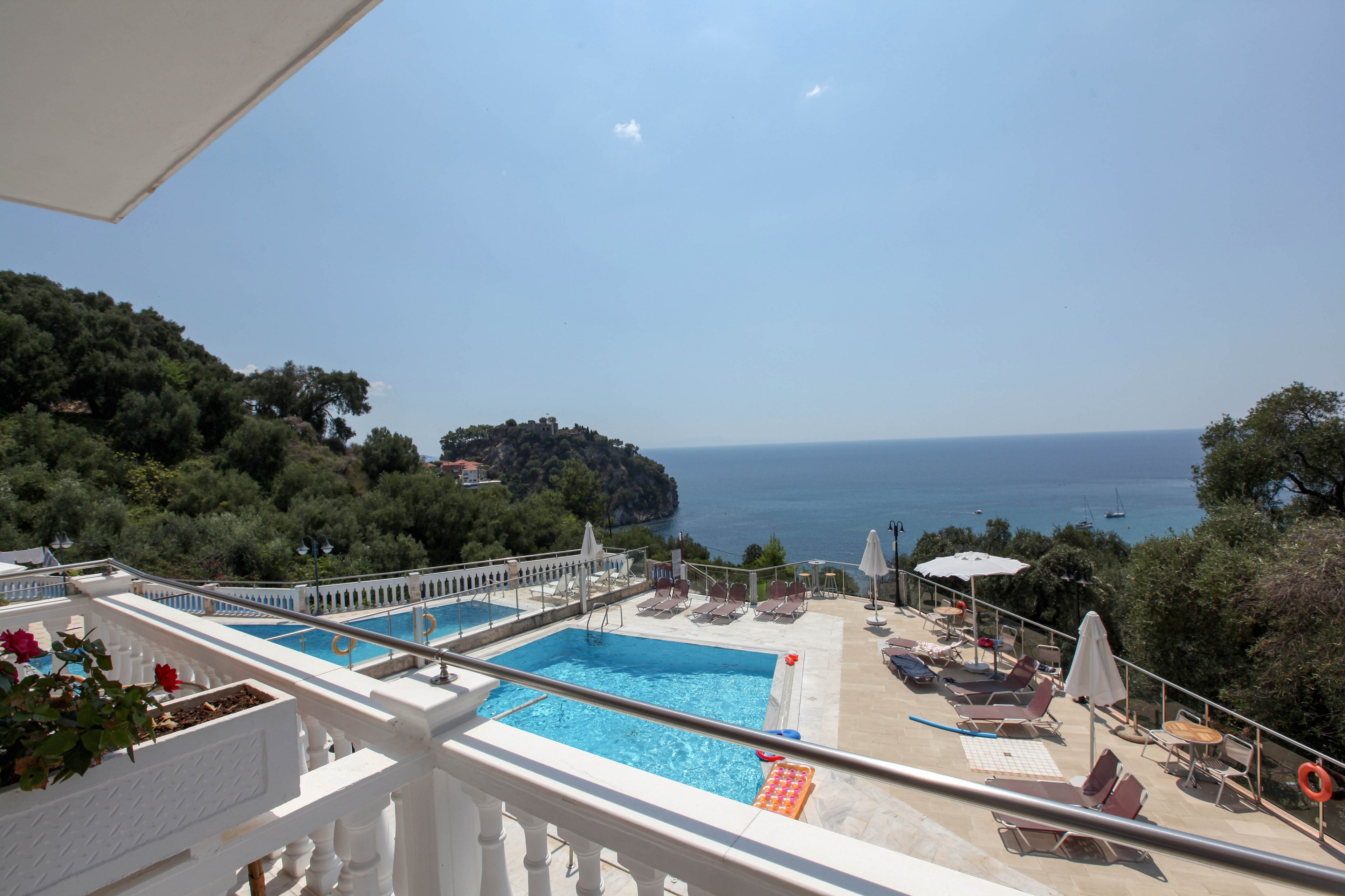 A spacious outdoor pool area with white railings overlooks a stunning blue ocean and lush green hills. Sun loungers and umbrellas are arranged around the pool, creating a relaxing vacation atmosphere. The scene is bathed in bright daylight, with clear skies and tranquil water. No visible people or text are present in the image. The overall mood is serene and inviting, perfect for leisure and relaxation.