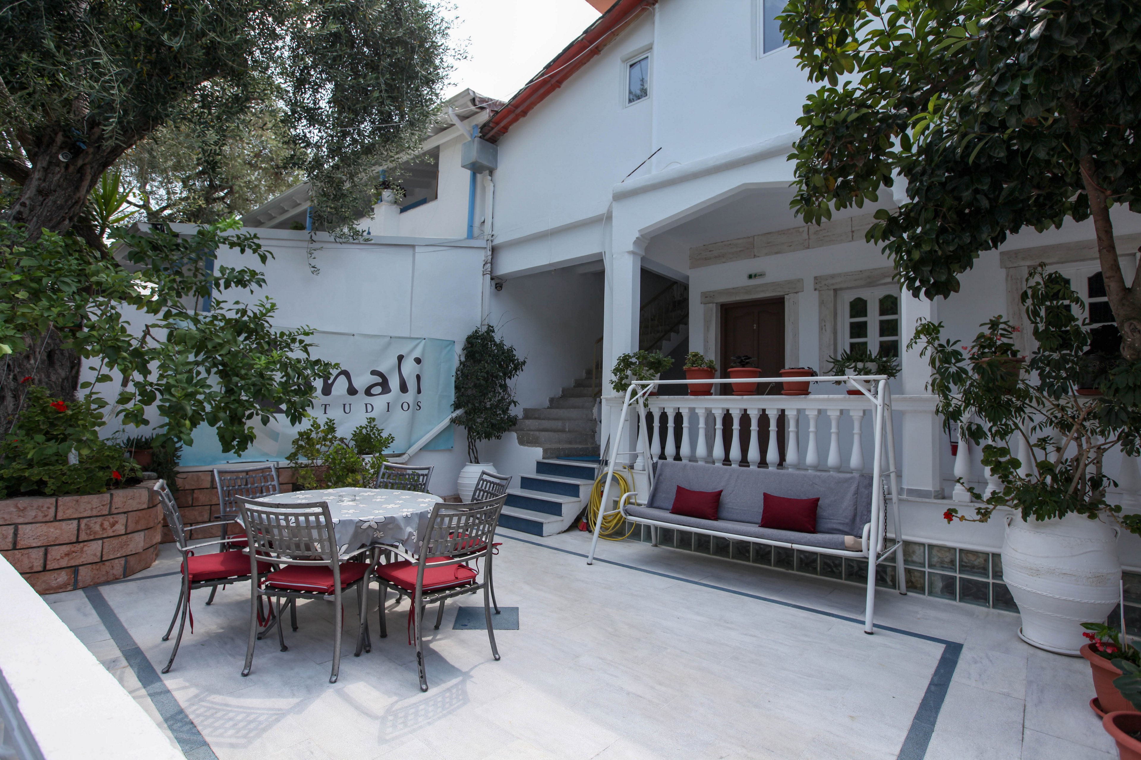 A bright outdoor patio features a metal table with six chairs, each with red cushions, set on a tiled floor. A white swing bench with colorful pillows sits under a covered porch, surrounded by lush green plants and trees. The setting is tranquil and inviting, ideal for gatherings or relaxation. The word 'nadi' is clearly visible on a sign in the background. The overall mood is peaceful and welcoming.