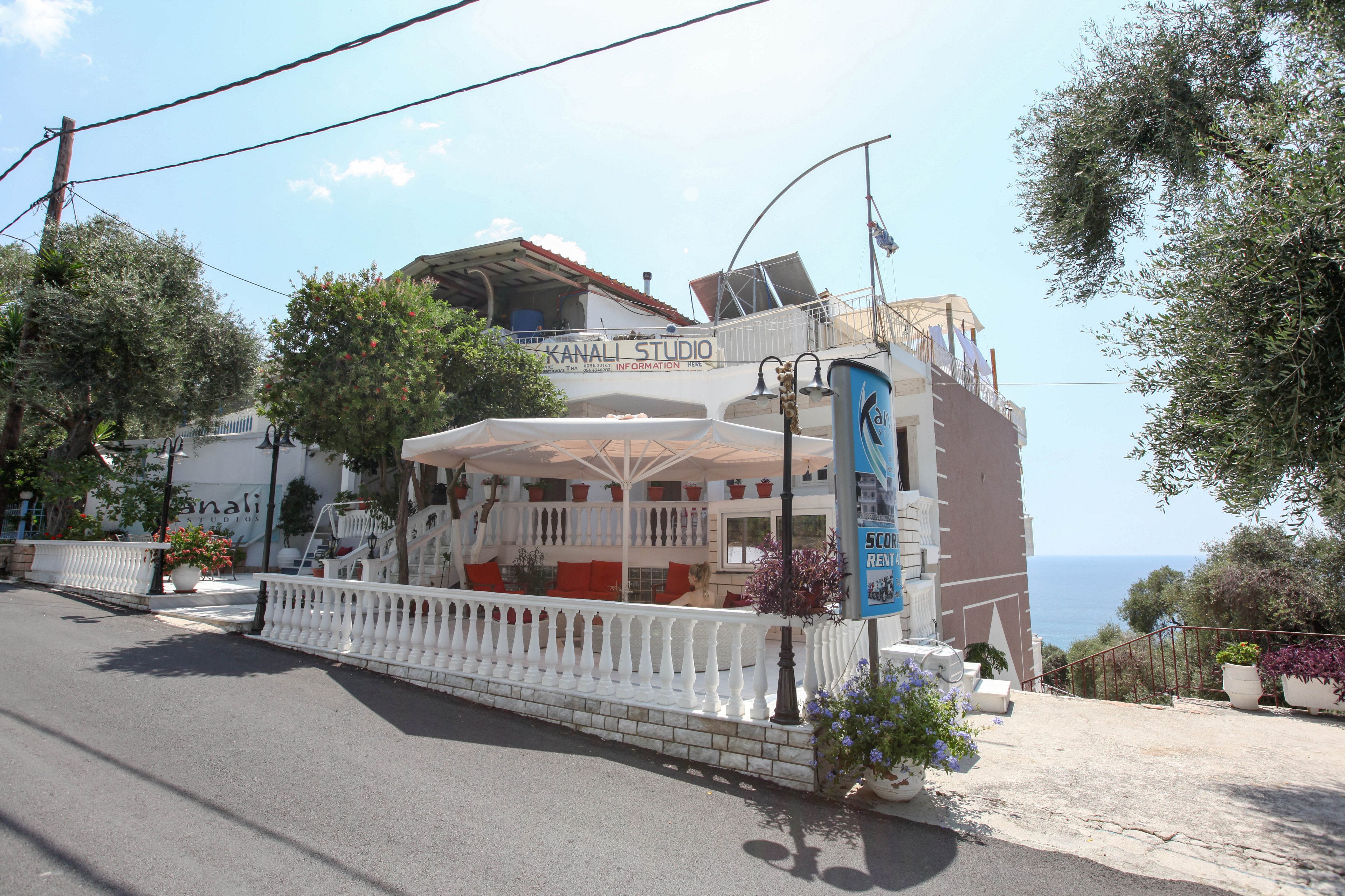 A white building labeled 'KANALI STUDIO' sits on a sloped street overlooking the sea. The scene features an outdoor terrace with white railings and shaded seating, surrounded by lush greenery and potted flowers. The setting is bright and sunny, with clear blue skies and a relaxed Mediterranean atmosphere. Visible signage includes the text 'KANALI STUDIO' and 'SEA VIEW.'