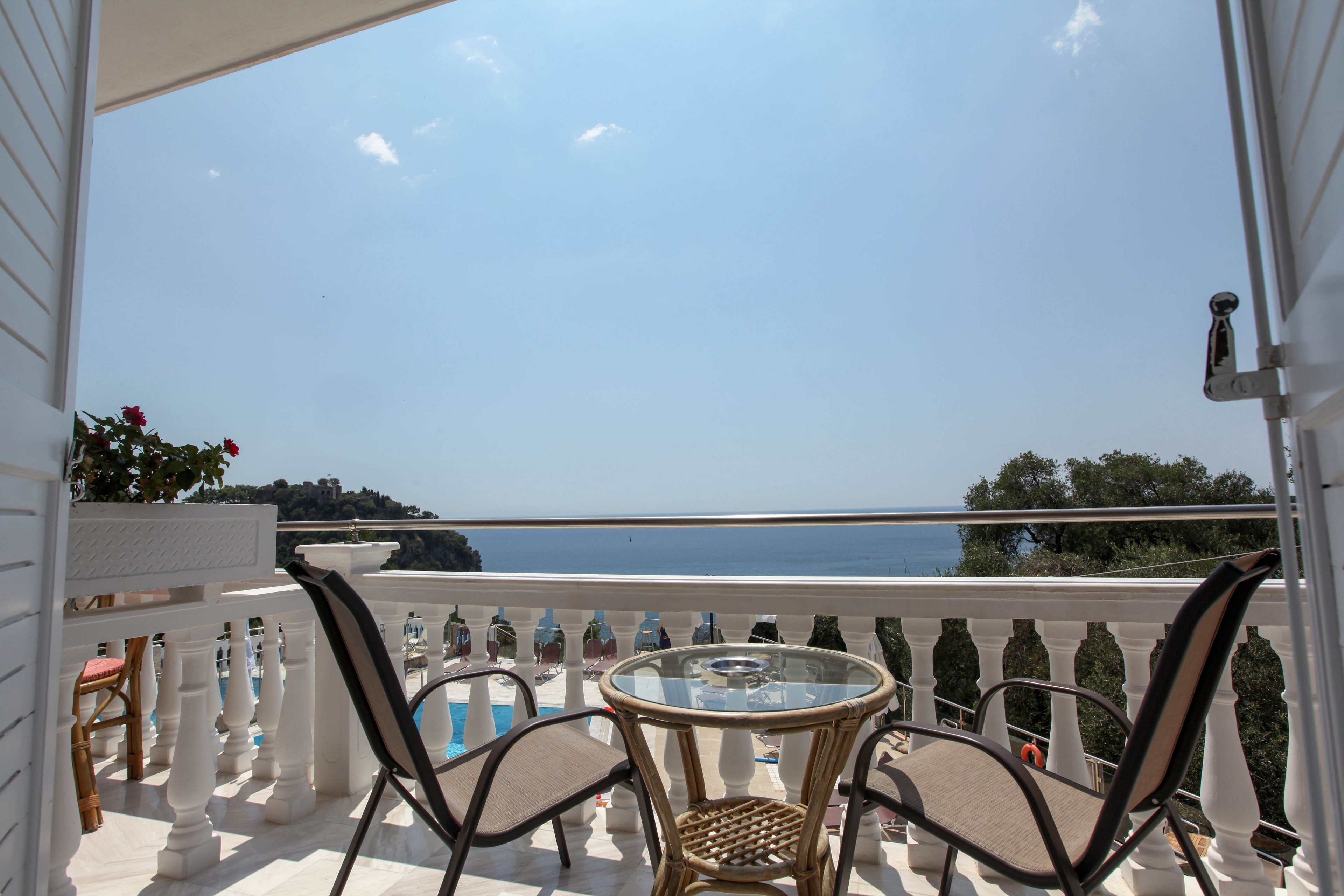 A bright balcony overlooks a scenic ocean view, featuring a small round glass table and two chairs. The setting is tranquil and sunlit, with white balustrades and potted plants adding to the relaxed atmosphere. The sky is clear, and the overall mood is peaceful and inviting, perfect for leisure or quiet conversation. No visible text or numbers are present in the image.
