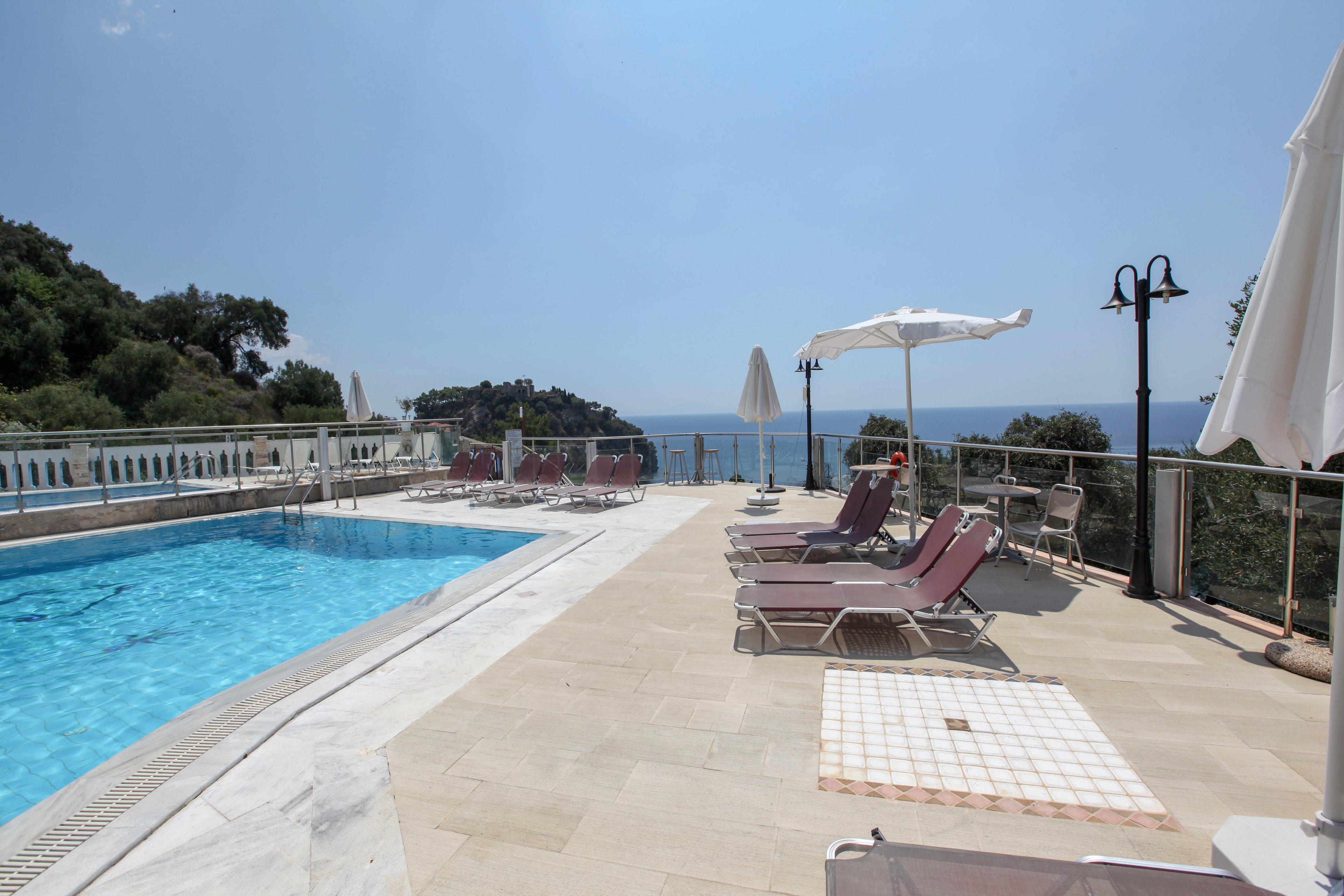 A serene outdoor swimming pool is set on a spacious terrace with several lounge chairs and umbrellas. The area overlooks a scenic ocean view, bordered by glass railings and surrounded by lush greenery. The setting is bright and tranquil, with clear skies and soft natural light, evoking a relaxed and luxurious vacation atmosphere. No people are present, emphasizing the peacefulness of the location.