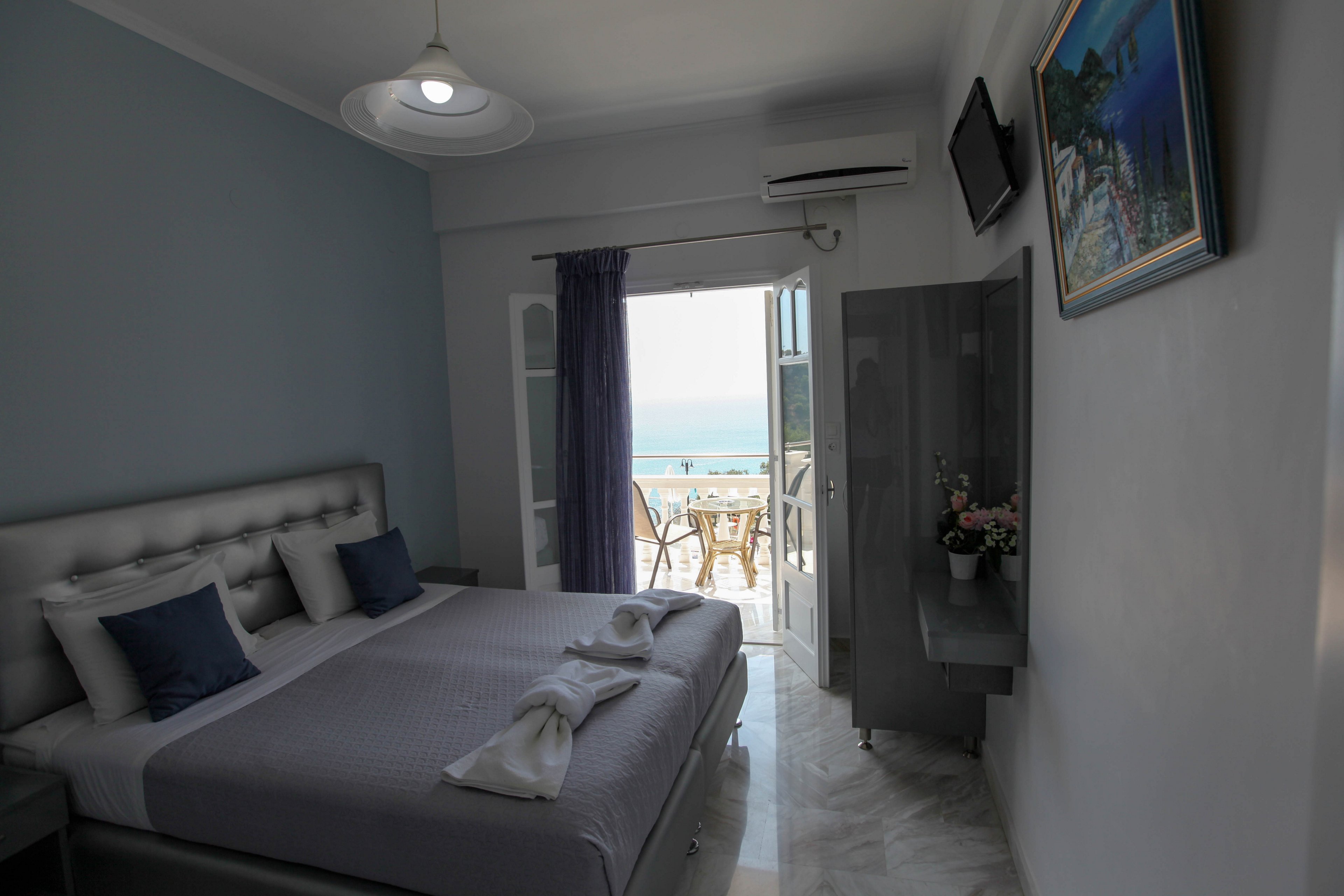 A contemporary hotel room features a neatly made double bed with gray and blue accents. The room opens to a balcony with a table and chairs, offering a scenic view of the sea. Soft natural light fills the space, highlighting minimalist decor, a wall-mounted TV, and a small flower arrangement. The overall mood is calm and inviting, with cool tones and a touch of elegance.