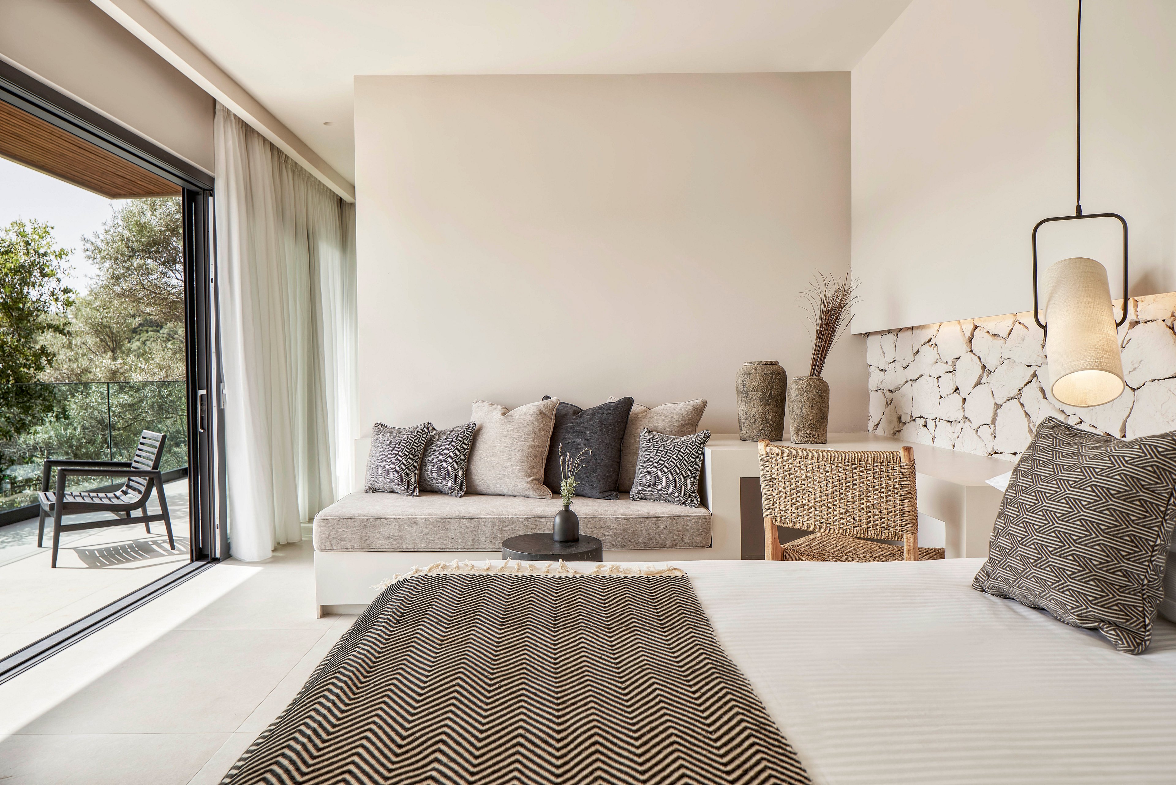 A contemporary bedroom featuring a neatly made bed with a patterned throw, a cozy seating area, and a workspace. The room is illuminated by natural light streaming through large glass doors that open to a balcony with greenery. Neutral tones and minimalist decor create a calm and inviting atmosphere.