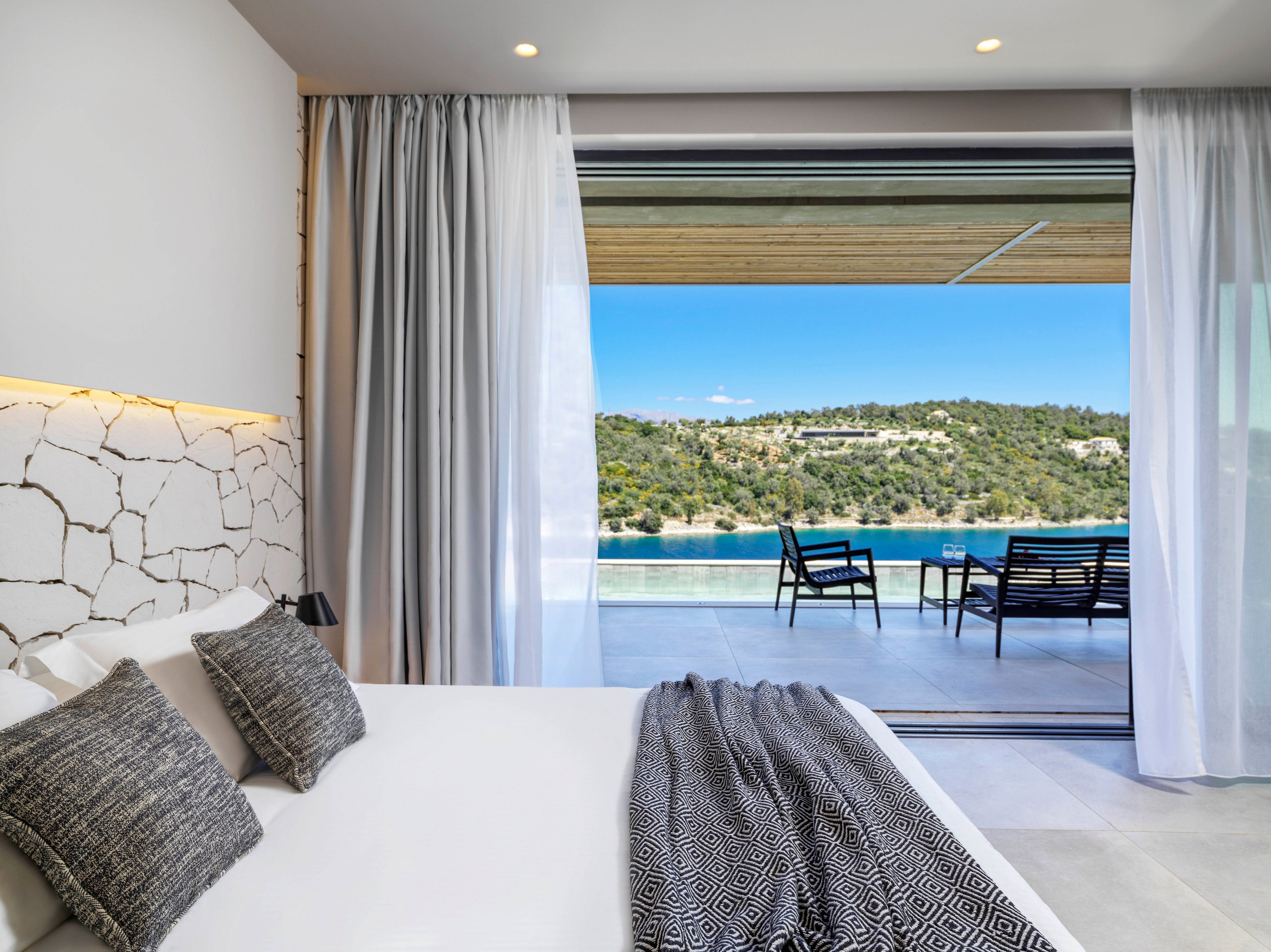 A contemporary bedroom featuring a white bed with patterned pillows and a textured blanket. The room opens to a spacious terrace with seating overlooking a serene pool and lush hillside scenery. Natural light enhances the minimalist design and tranquil atmosphere.