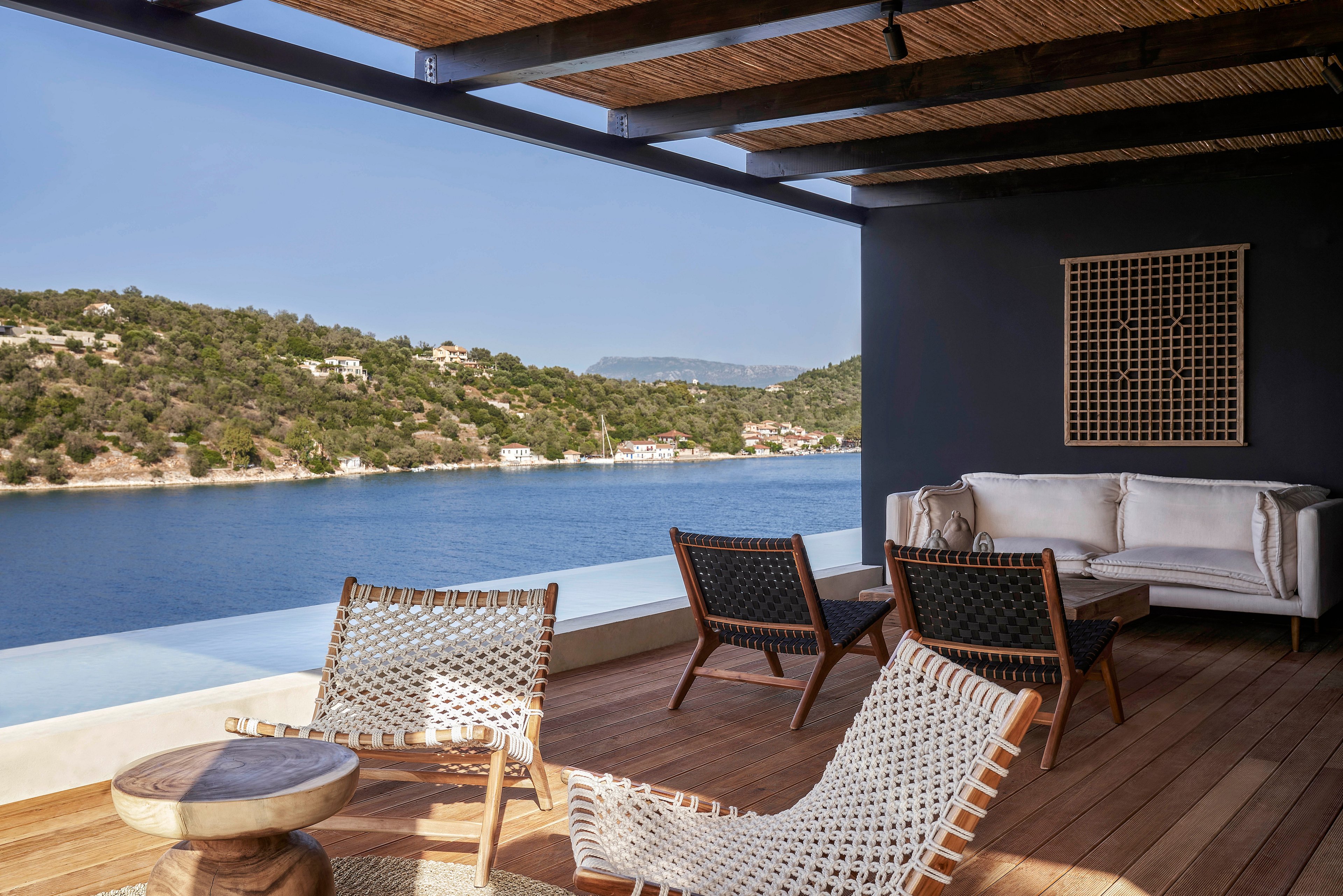 A stylish outdoor terrace featuring woven chairs, a wooden table, and a comfortable sofa overlooks a calm waterfront. The setting is enhanced by a wooden deck and a shaded pergola, offering a relaxing ambiance. The view includes lush green hills and clear blue water under a sunny sky.