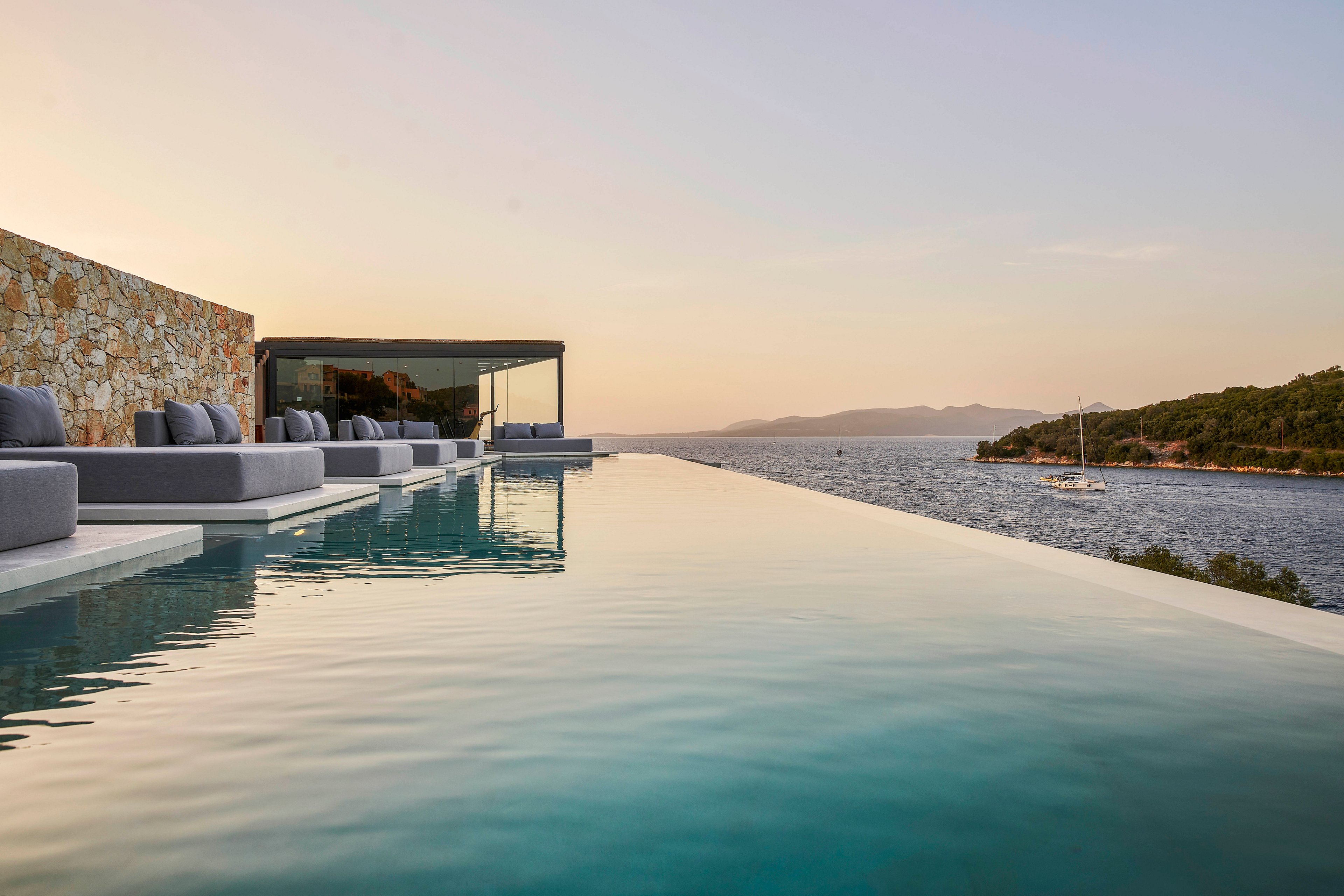 A luxurious infinity pool stretches towards a tranquil coastal horizon at sunset. The setting features modern lounge seating and a sleek architectural design. The calm water reflects the warm hues of the sky, creating a peaceful ambiance. A distant sailboat and lush greenery enhance the scenic beauty.