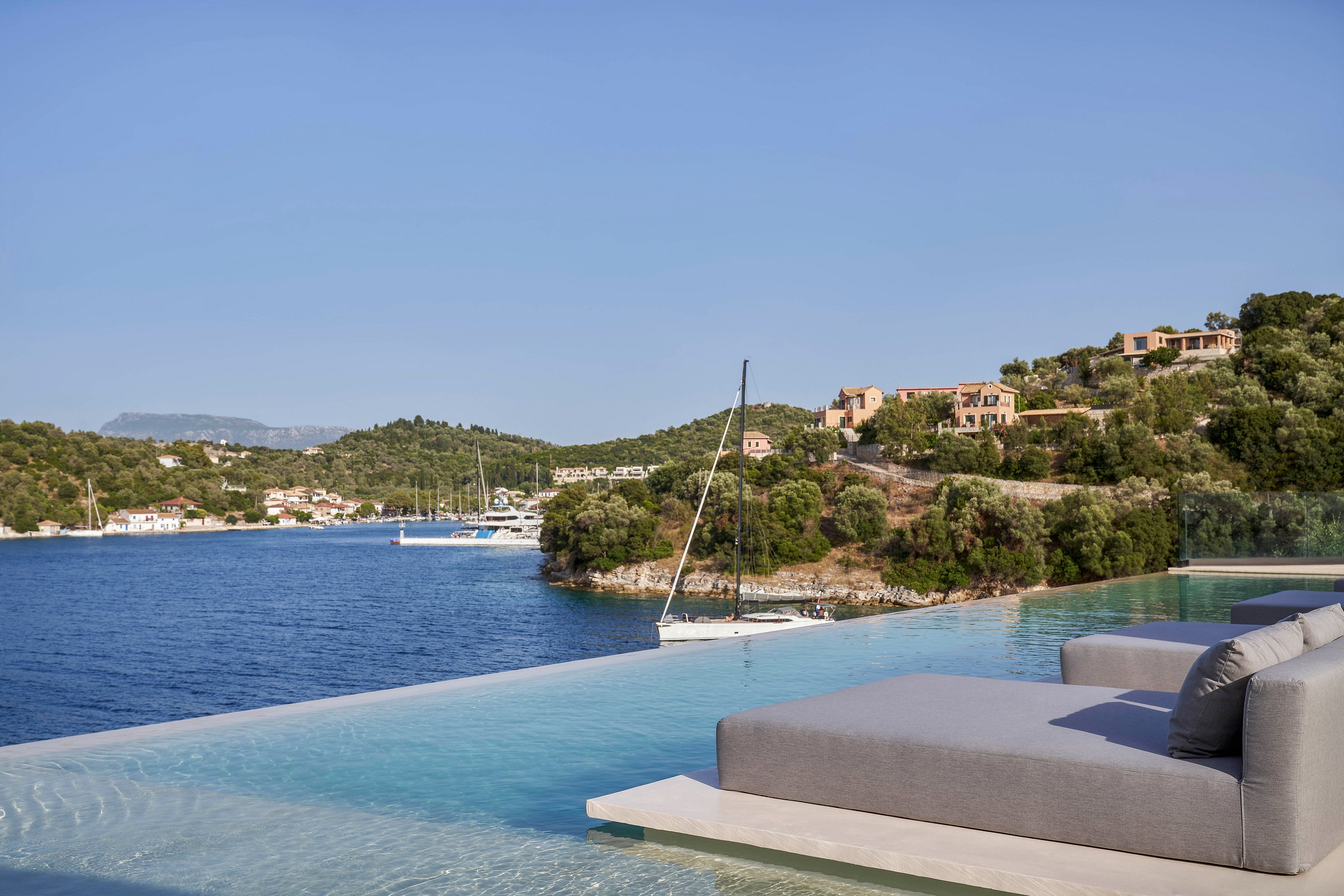 A luxurious infinity pool with modern gray lounge chairs overlooks a tranquil coastal bay. The setting features calm blue waters, a sailboat, and lush green hills dotted with villas. The clear sky enhances the peaceful and scenic atmosphere.
