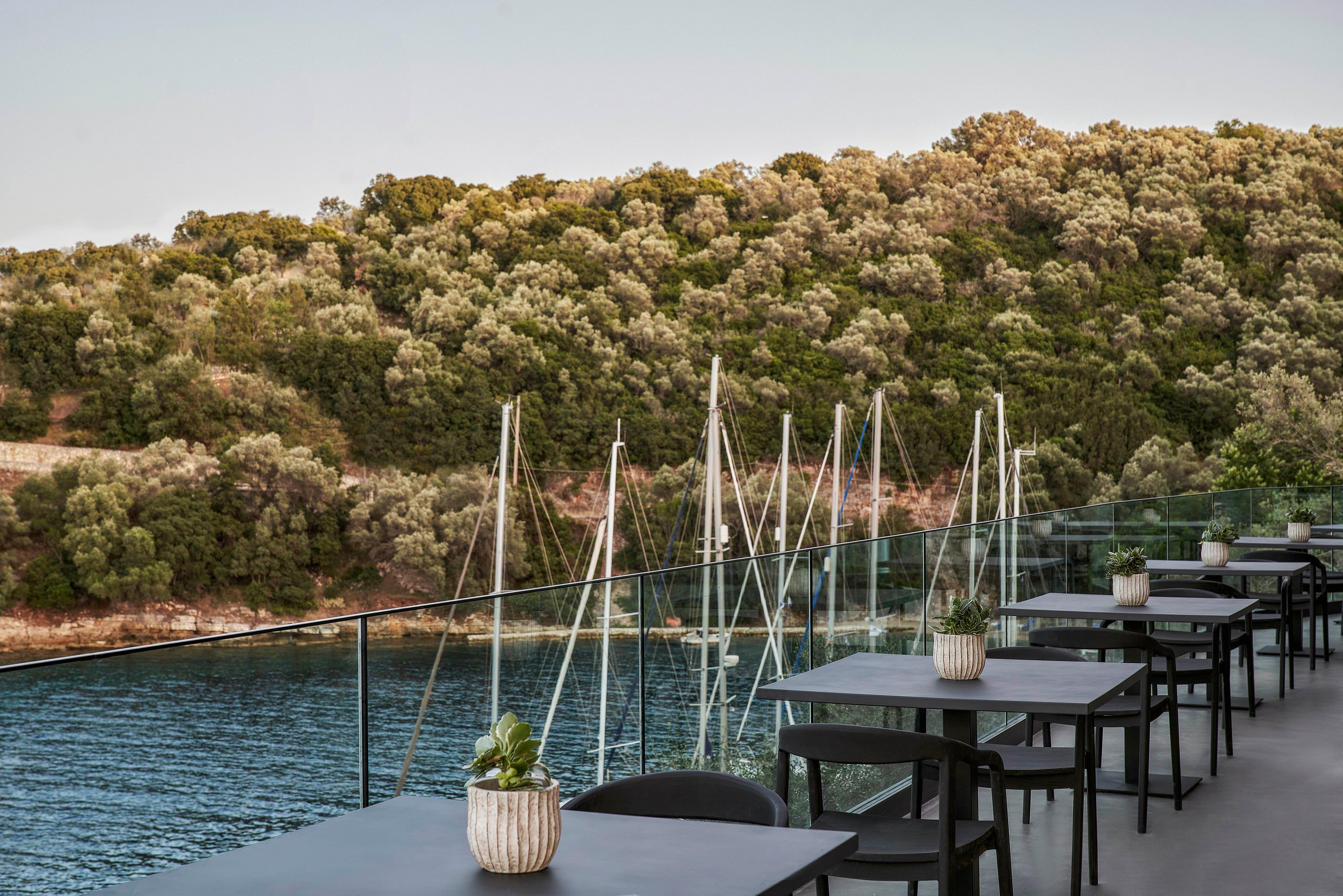 An outdoor dining area overlooking a serene waterfront with docked sailboats. The setting features modern black tables and chairs, complemented by potted plants. The backdrop includes lush green hills and calm waters, creating a tranquil atmosphere.