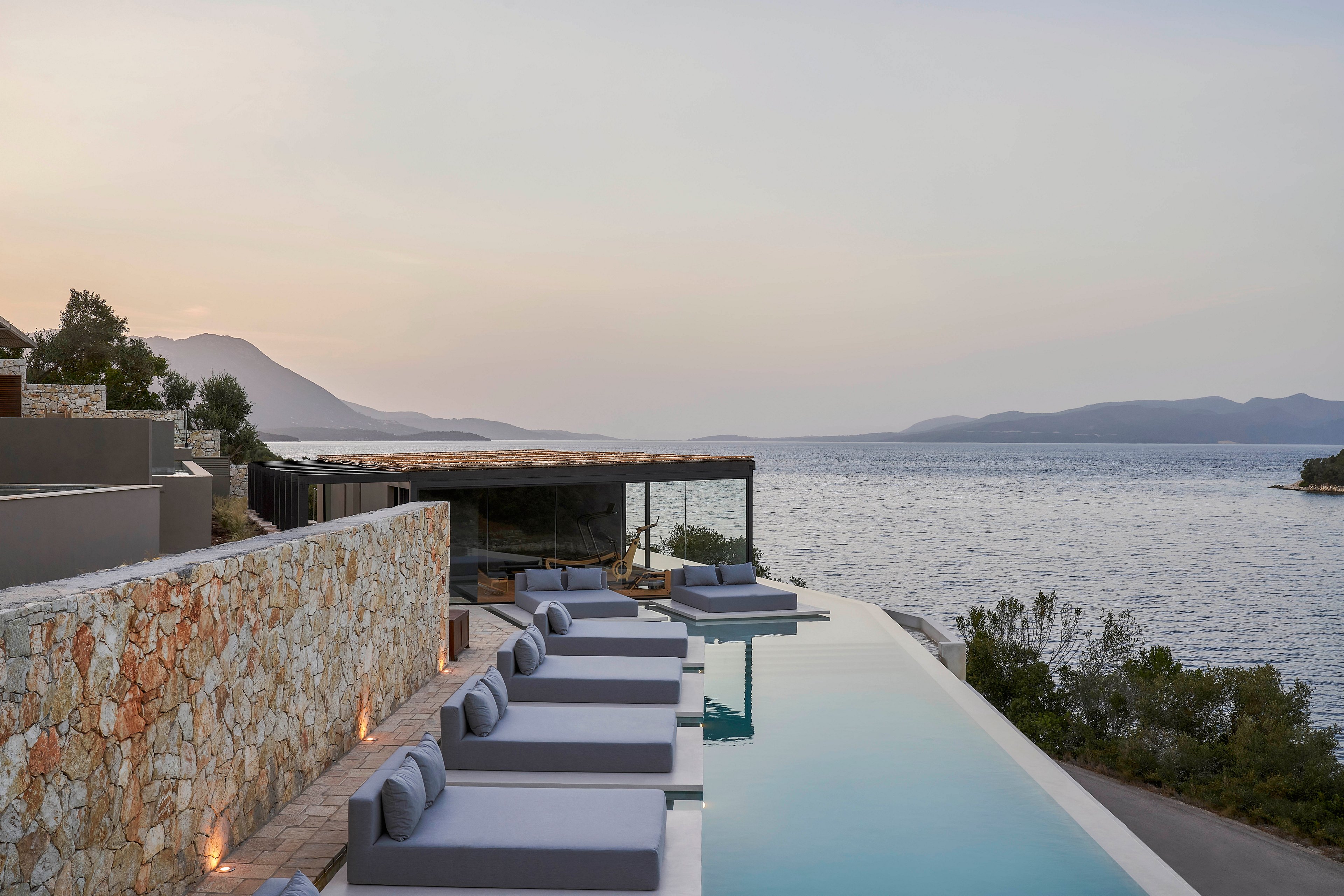 A tranquil infinity pool with modern lounge chairs overlooks a calm sea and distant mountains at sunset. The setting features a stone wall and contemporary architecture, creating a luxurious and serene atmosphere. The soft lighting enhances the peaceful ambiance of the outdoor space.