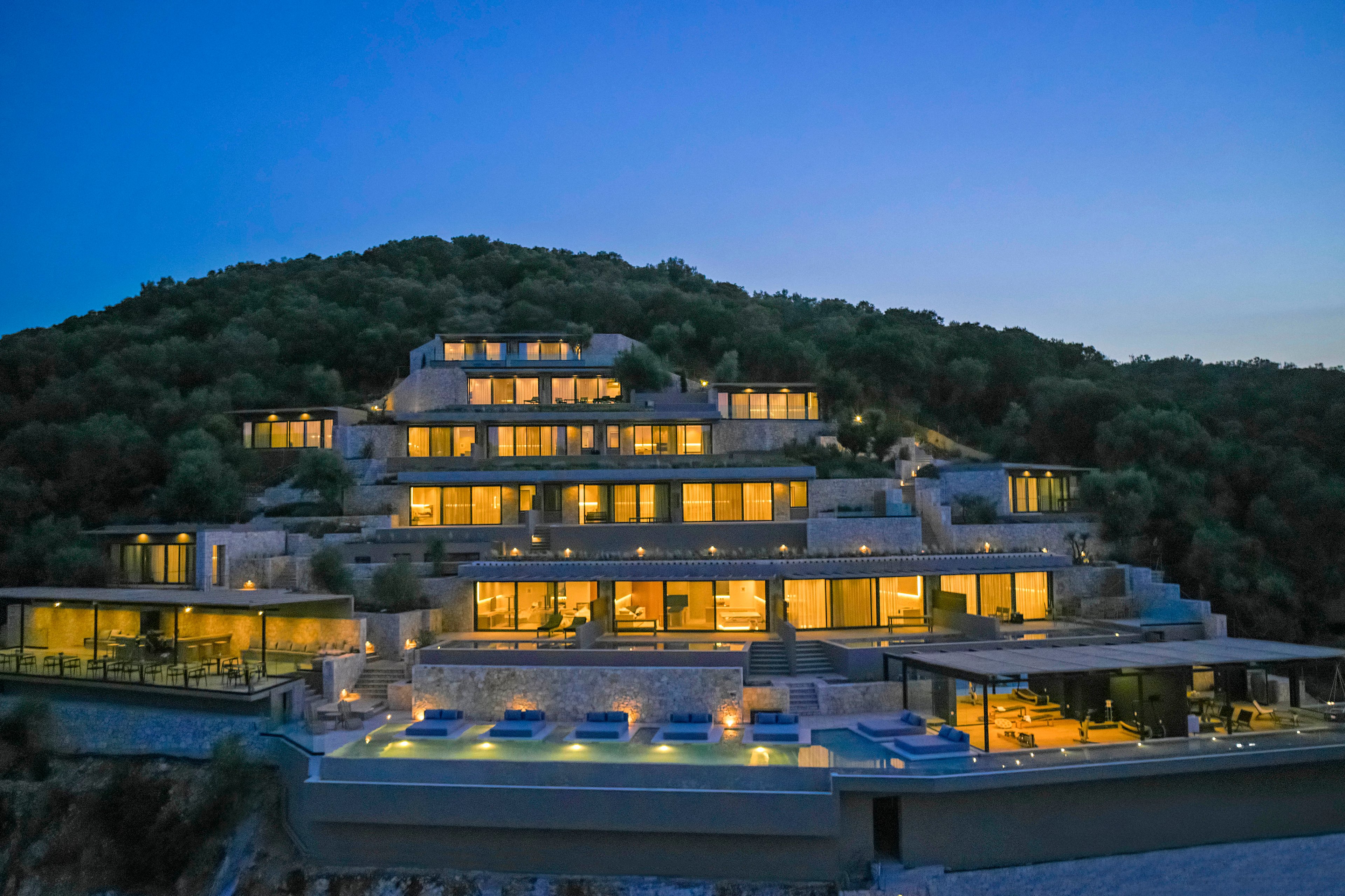 A multi-tiered luxury villa is illuminated against a twilight sky, nestled on a lush hillside. The modern architecture features expansive glass windows and terraces, with a series of pools and outdoor seating areas. The serene setting highlights the villa's grandeur and tranquil ambiance.