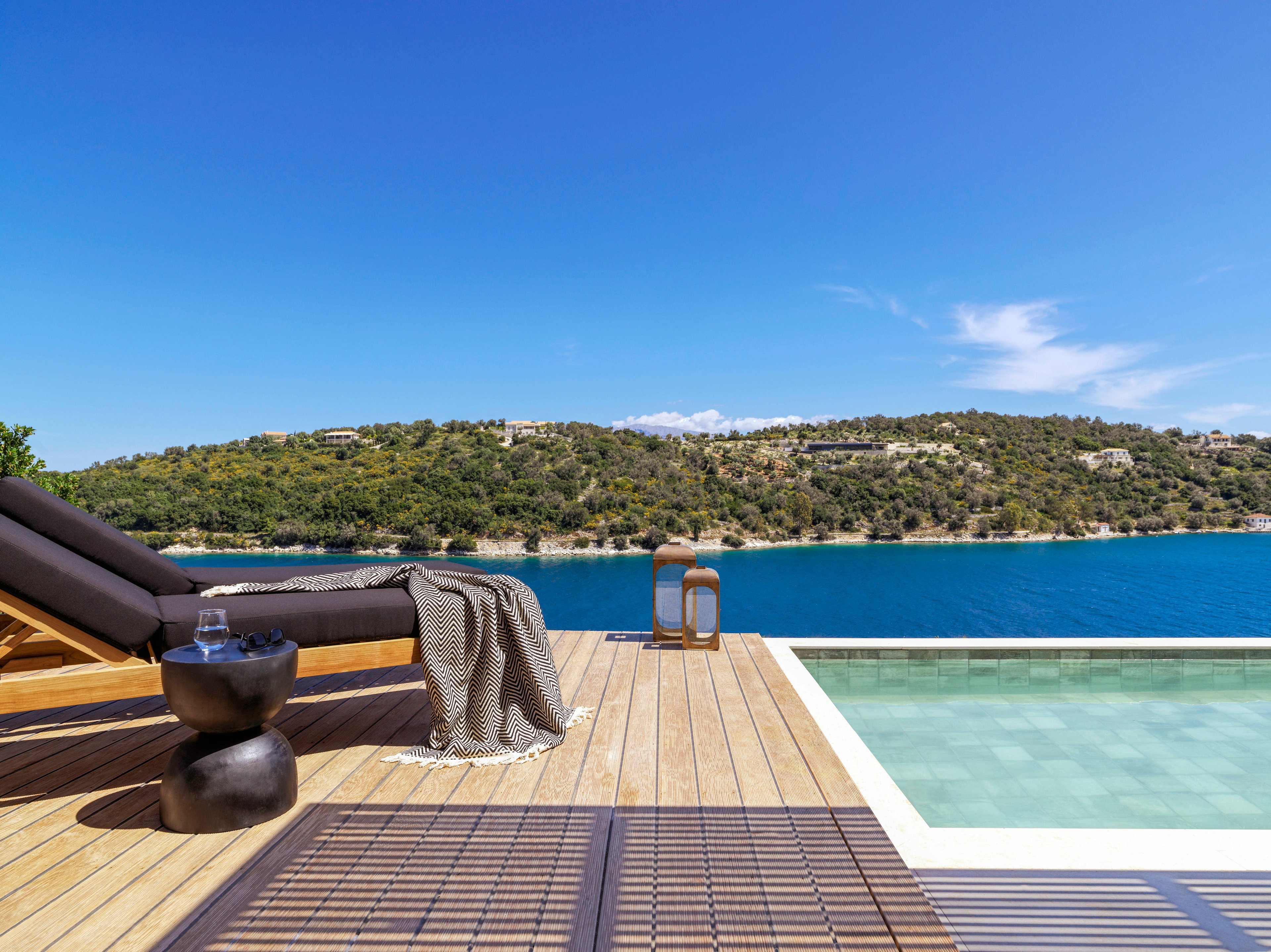 A modern poolside deck featuring a black lounge chair with a textured blanket draped over it. The setting overlooks a serene blue bay surrounded by lush green hills under a clear blue sky. The wooden deck includes a small black side table and a pool with calm, reflective water.